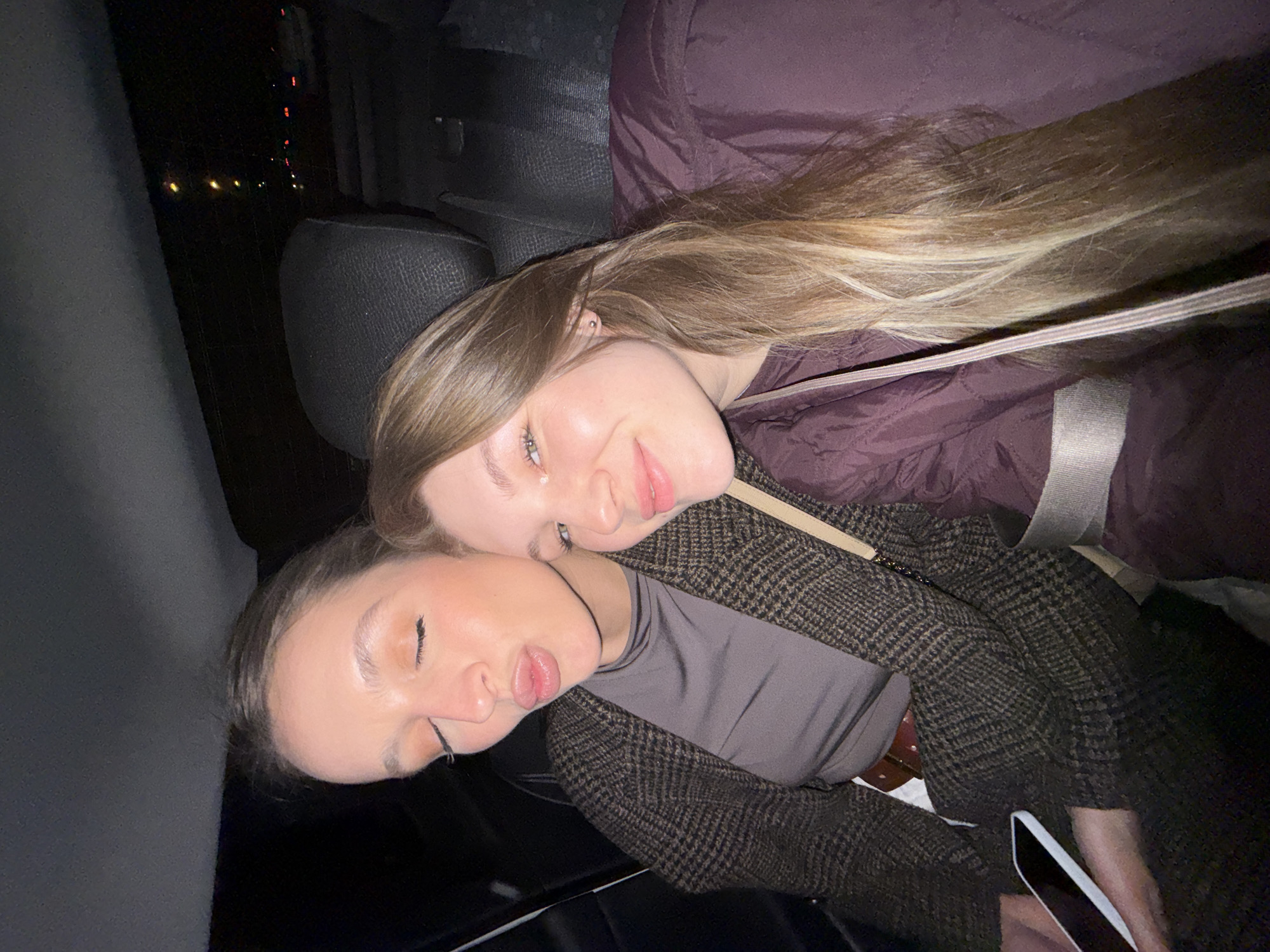 Two young women are in what appears to be a car, possibly at night. One woman with blonde hair is awake and looking at the camera, wearing a maroon jacket and a seatbelt. The other woman with darker hair is asleep with her head resting on the shoulder of the first woman, wearing a patterned blazer and a gray shirt. Part of a car seat is visible in the background. There's no indication that the scene is in Vozdovac Urban Municipality, Serbia.
