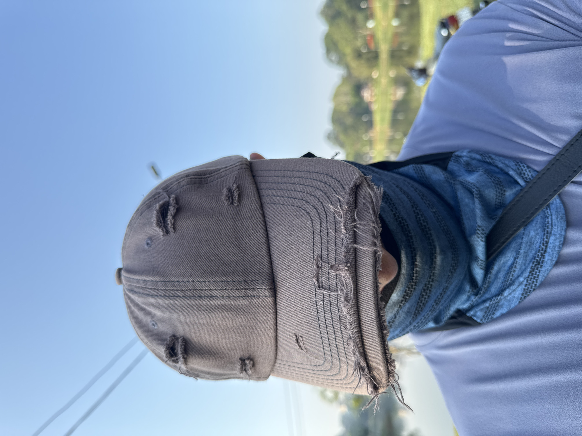The image, taken from a low angle, shows a person wearing a grey, distressed baseball cap and a blue patterned face covering. They are wearing a white shirt and a black strap is visible across their shoulder. The background is blurry but appears to be a landscape with a mix of vegetation and possibly buildings under a clear blue sky. The image's tilt suggests it was taken casually. It is hard to definitively identify any exact location.