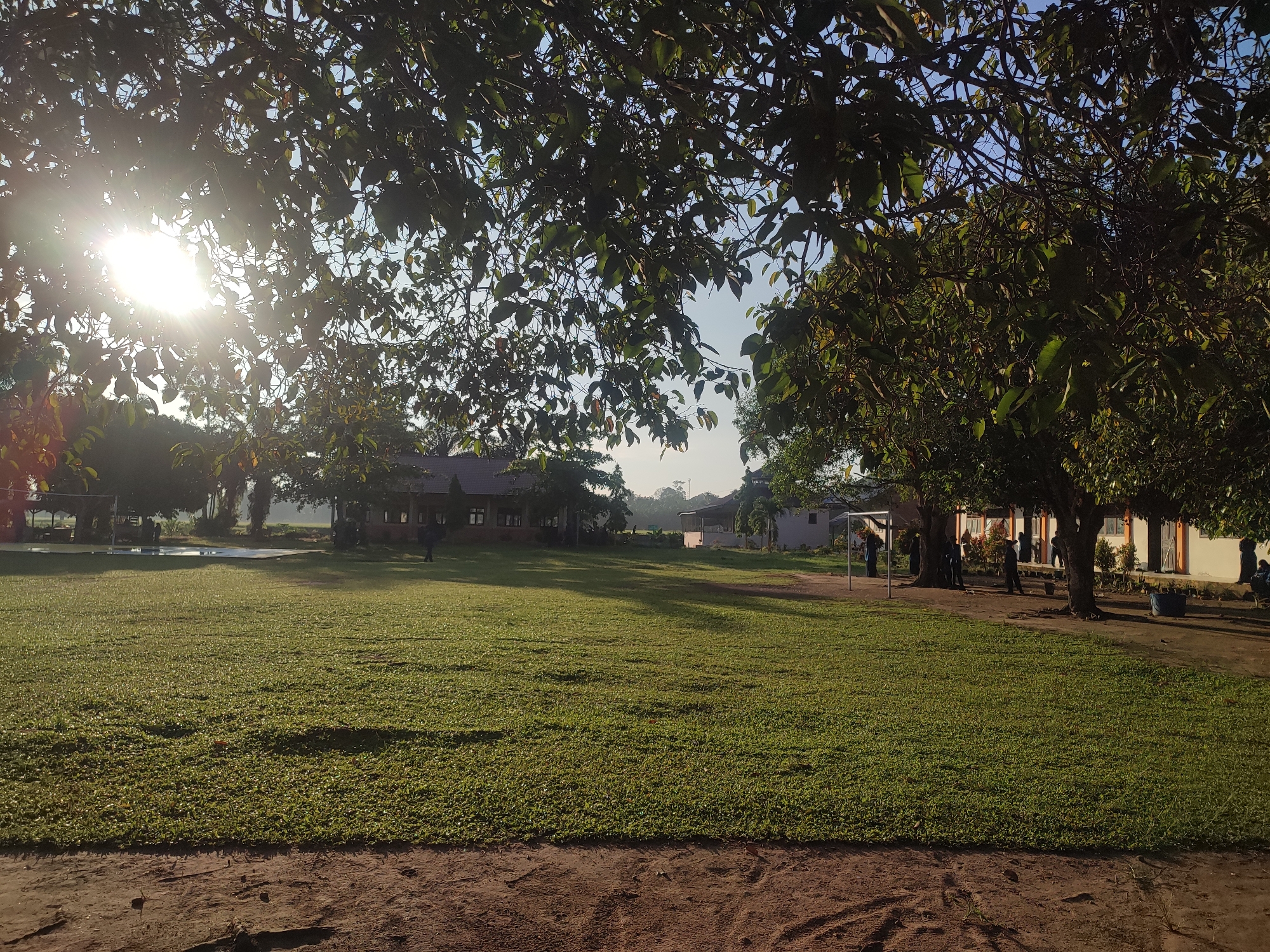 The image shows a grassy field in Blangriek, Indonesia. Buildings are visible in the background, partially obscured by trees. The sun shines brightly through the leaves of trees in the upper left corner of the frame, creating a strong contrast. Several people are near one of the buildings on the right side of the image. The foreground consists of bare earth.