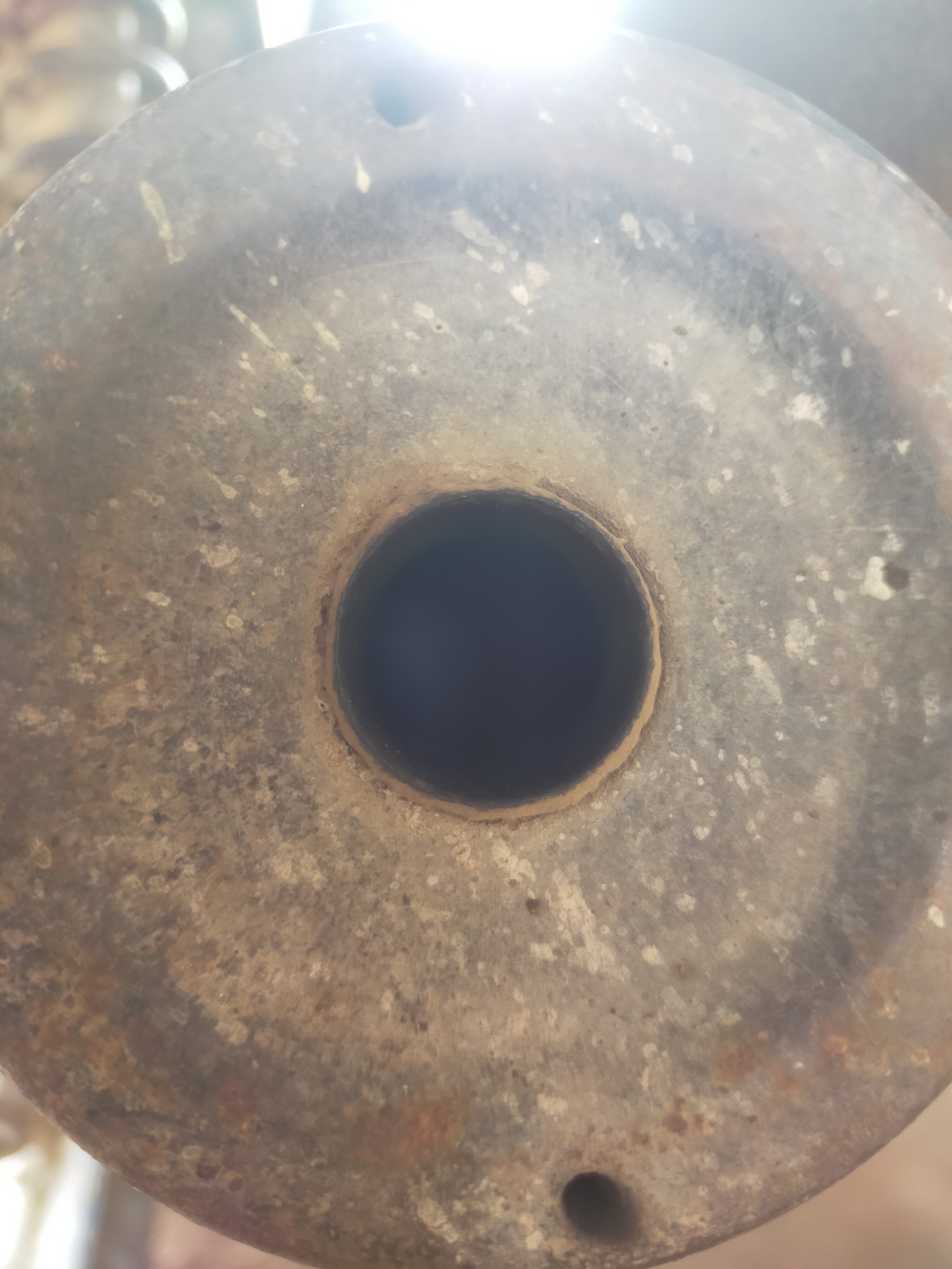 The image shows a close-up, overhead view of a rusty, circular metal object with a hole in the center. The surface of the object is textured and discolored, with patches of rust and what appear to be white or light-colored speckles. There is a bright light source visible at the top of the image, causing glare and overexposure in that area. The hole in the center is dark, obscuring whatever might be behind it. The overall impression is of an aged and weathered industrial or mechanical component. The location information provided is irrelevant to the visual content of the image.