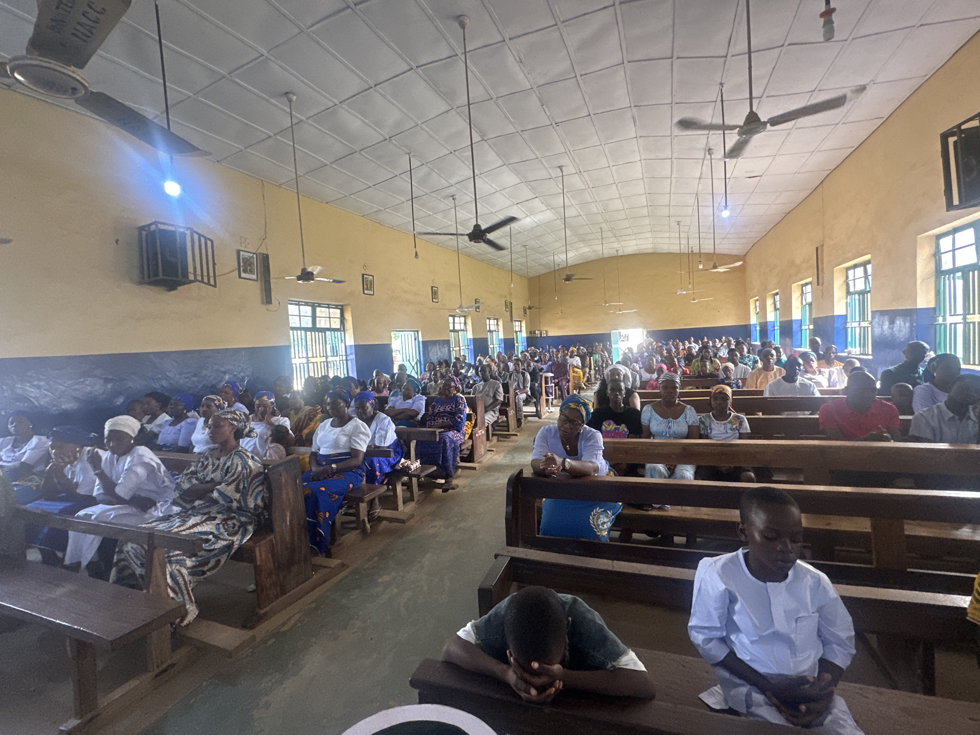 The image depicts the interior of a church in Akwanga, Nigeria, filled with a congregation seated on wooden benches. The room has a high ceiling with multiple ceiling fans, and the walls are painted yellow with a blue stripe near the floor. Natural light streams in from windows along the side of the building. In the foreground, several people are visible, including a young boy in a white garment with his hands clasped. Further back, a man is bowed with his head resting on his hands. The congregation consists of men, women, and children, many of whom are wearing traditional African attire. The overall atmosphere appears to be one of prayer or worship.