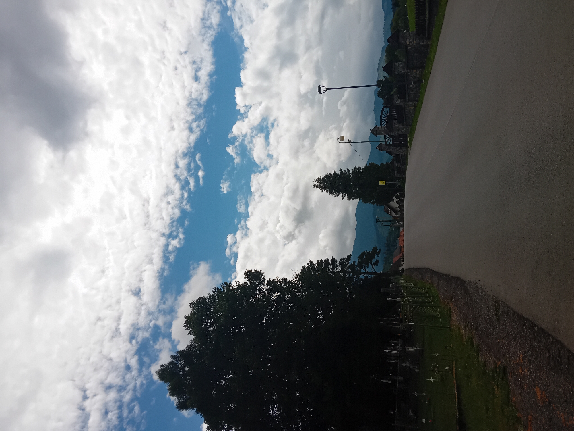 The image depicts a road leading through a village in Zuberec, Slovakia. A dark, paved road dominates the foreground, curving slightly to the right. To the left of the road, a fence encloses a grassy area with numerous crosses, indicating a cemetery. A large, dark silhouette of a tree stands prominently on the left side. Further into the scene, a village can be seen with houses and a large, dark green fir tree. A stone wall with a decorative gate is visible on the right side of the road, bordering a field. Two streetlights are present, one with a contemporary design and another older, spherical one. The background features rolling hills and mountains. The sky above is partly cloudy, with patches of blue sky visible between dramatic white and gray clouds. No people are visible in the image.
