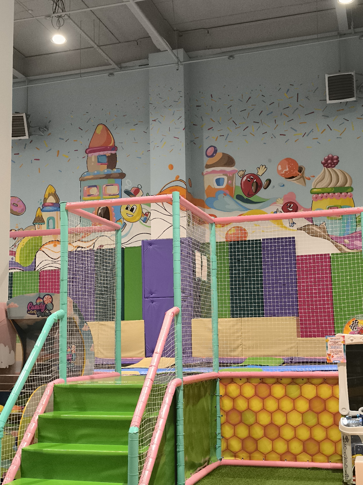 The image depicts the interior of a children's indoor playground, likely a "Candy Activity Park" as indicated by signage. The environment is brightly colored and features a candy-themed mural on the wall, including depictions of candy houses, ice cream cones, and sprinkles. The playground equipment includes a multi-level structure with green steps, pink and turquoise safety railings, and white netting. Various padded sections in shades of purple, green, and pink are visible within the play structure. A portion of a white play-themed easel or whiteboard is present in the lower right corner. The lighting appears to be artificial, originating from ceiling fixtures. No people are visible in the image. The scene is set indoors, likely in a commercial establishment, and there is no specific visual information to pinpoint its location within the city of Qarağandı city administration, Kazakhstan.