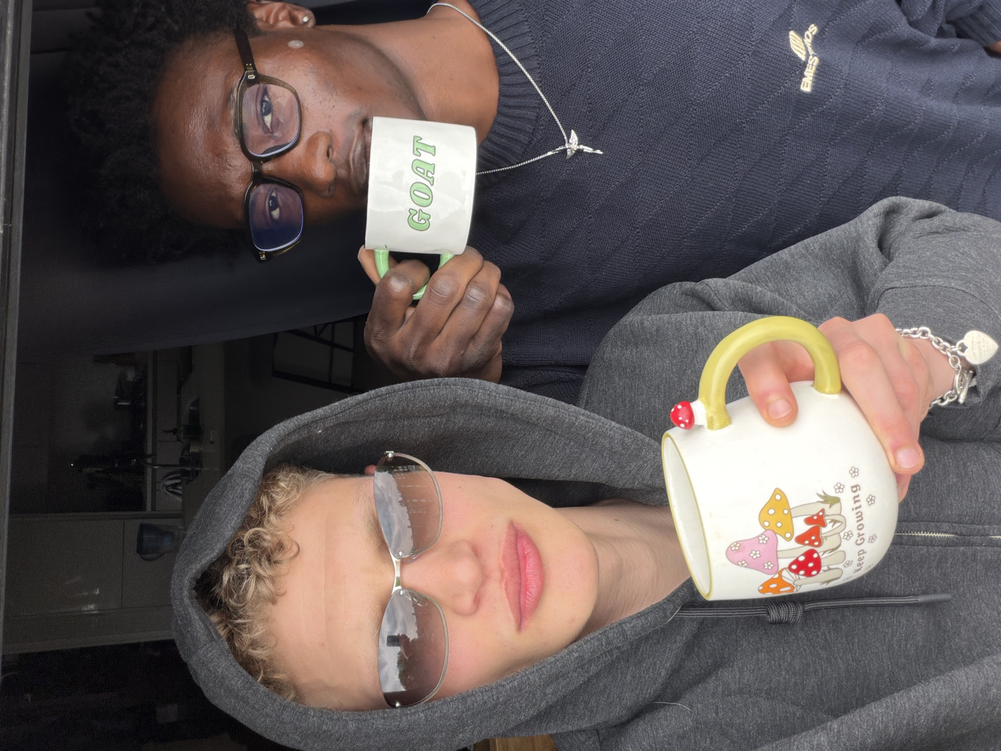 Two individuals are depicted in the image, oriented in opposite directions. The person at the top, of darker complexion, is wearing eyeglasses and a dark, textured sweater, holding a white mug with the word "GOAT" printed in green. A silver necklace with a charm is visible around their neck.
The person at the bottom, of lighter complexion, is wearing a gray hoodie and reflective sunglasses. They are holding a white mug adorned with cartoonish mushrooms and the phrase "Keep Growing." A silver bracelet with a heart-shaped charm is on their wrist.
The background is blurred and indistinct, but suggests an indoor environment, potentially within a building or structure. The specific location in London, United Kingdom, cannot be determined from the provided image.
