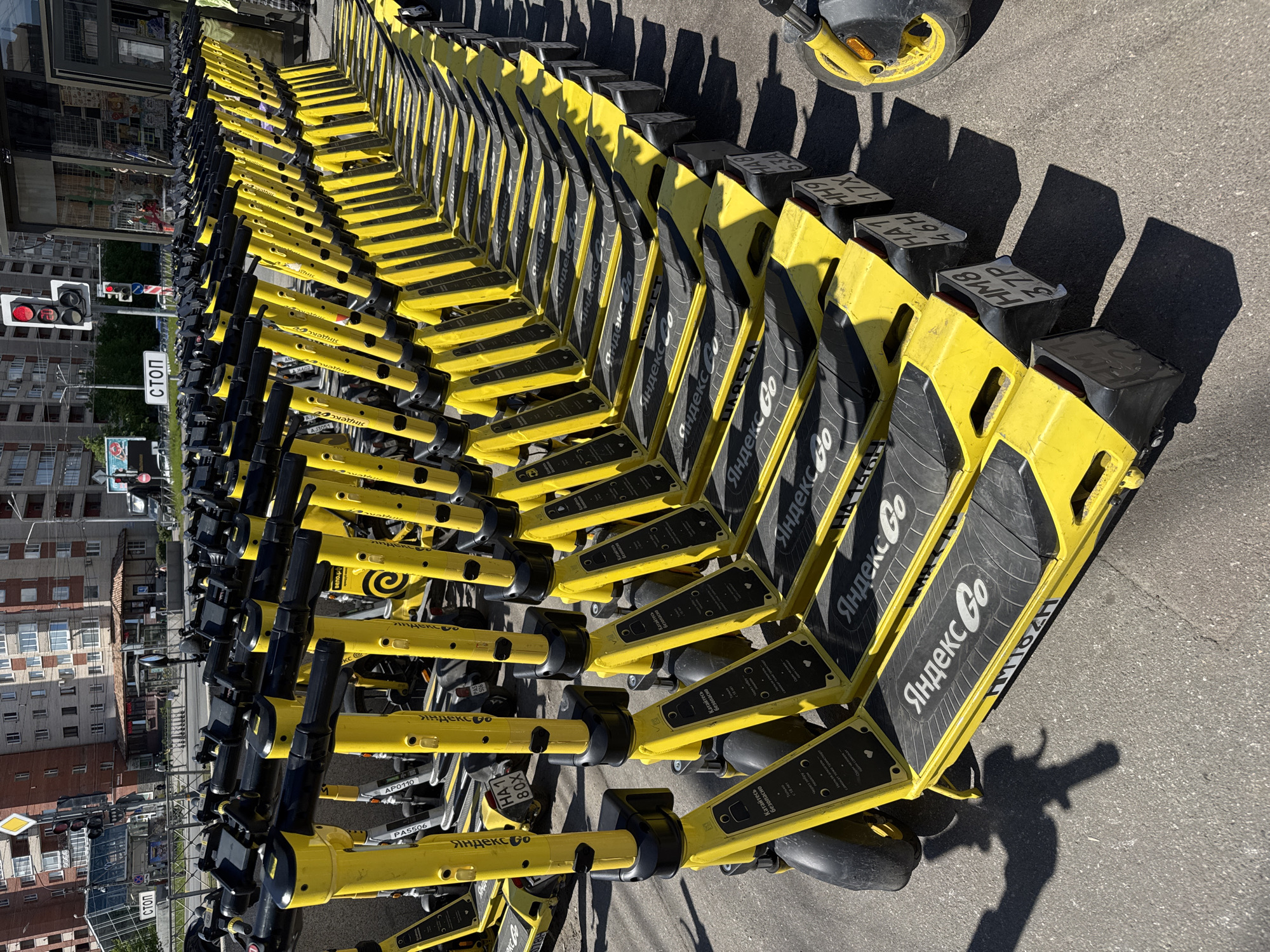 The image displays a large number of bright yellow electric scooters, primarily branded "Яндекс Go" (Yandex Go) and "Яндекс" (Yandex), parked in multiple orderly rows on an asphalt street. The scooters feature contrasting black handlebars, grips, and footboards. Many scooter decks show the Cyrillic text "Катайтесь безопасно" (Ride safely), indicating safety instructions. Various alphanumeric identifiers like "PA5506," "AP0110," "HA1 80X," "HA1 46H," "37P," "OZET," and "TMTOH" are visible on different parts of the scooters. One scooter also displays "вело.рф."
In the background, a typical urban landscape unfolds with multi-story buildings. A red traffic light and a street sign reading "СТОП" (STOP) in Cyrillic are prominent on the left side of the frame. The scene is illuminated by bright sunlight, casting long, distinct shadows from the scooters onto the pavement, suggesting it is mid-day or afternoon on a clear, sunny day. The overall setting, with its Cyrillic text and city architecture, is consistent with Saint Petersburg, Russia, showcasing a common urban shared mobility service. The scooters are stationary, seemingly available for public use.