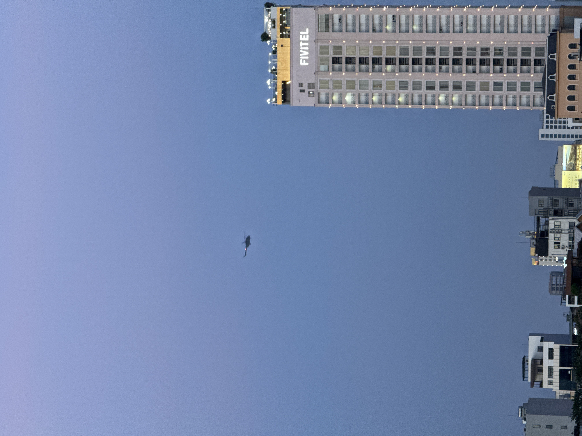 A tall, modern skyscraper with the name "FIVITEL" illuminated in white lettering dominates the right side of the frame. The building is lit with numerous small, bright lights along its facade and at the top, suggesting it is evening or night. The skyscraper appears to be located in the city of Da Nang, Vietnam. In the sky, a dark silhouette of a helicopter is visible in flight. Below the skyscraper, other buildings of varying heights and architectural styles are visible, indicating an urban setting. The sky is a gradient of twilight colors, transitioning from a pale lavender at the top to a deep blue towards the horizon. The scene captures a portion of the Da Nang skyline during dusk, with the FIVITEL building serving as a prominent landmark.