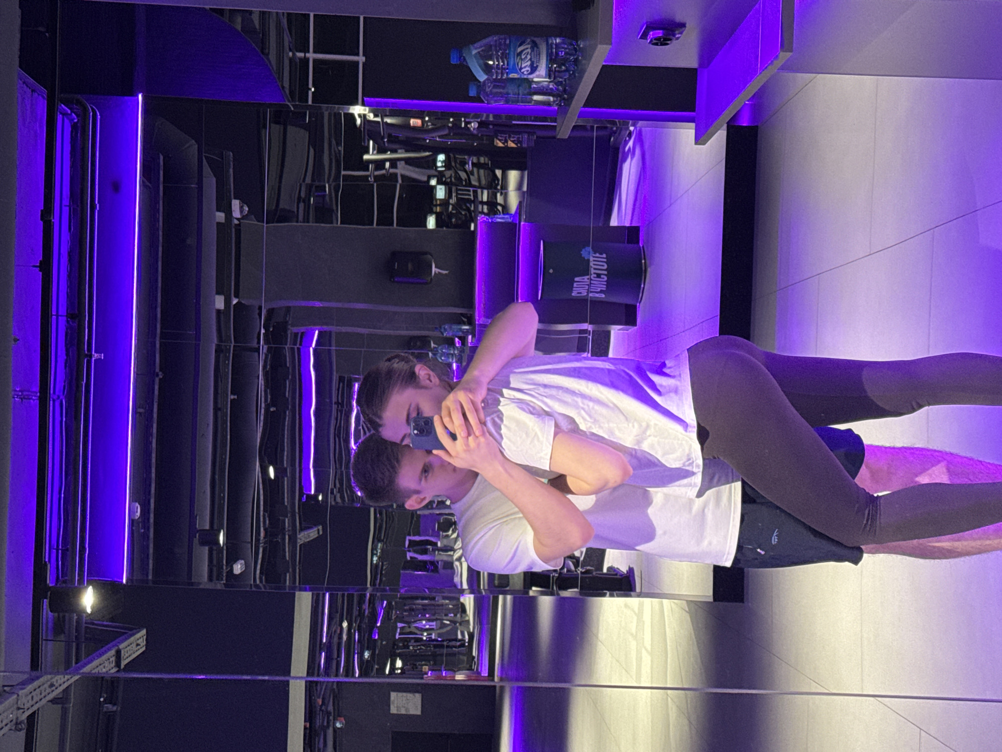 The image depicts a young couple taking a mirror selfie in a gym. The man, on the left, is wearing a white t-shirt and dark shorts. He has his arm around the woman, who is on the right. She is wearing a white t-shirt and dark leggings, and is holding a smartphone to take the photo. They are standing close together, with their heads tilted towards each other.
The gym is modern and dimly lit, with purple LED lighting illuminating the space. Large mirrors line the walls, reflecting the gym equipment and the couple. In the background, exercise machines are visible, as well as water bottles on a shelf and a trash can with Cyrillic lettering that reads "СИЛА В ЧИСТОТЕ" (Strength in Cleanliness). The floor is tiled and reflects the purple light.
The specific location is a gym in Krasnoyarsk, Russia. The presence of Cyrillic text on the trash can strongly suggests a Russian-speaking environment, and the gym's modern aesthetic is consistent with facilities found in urban centers.