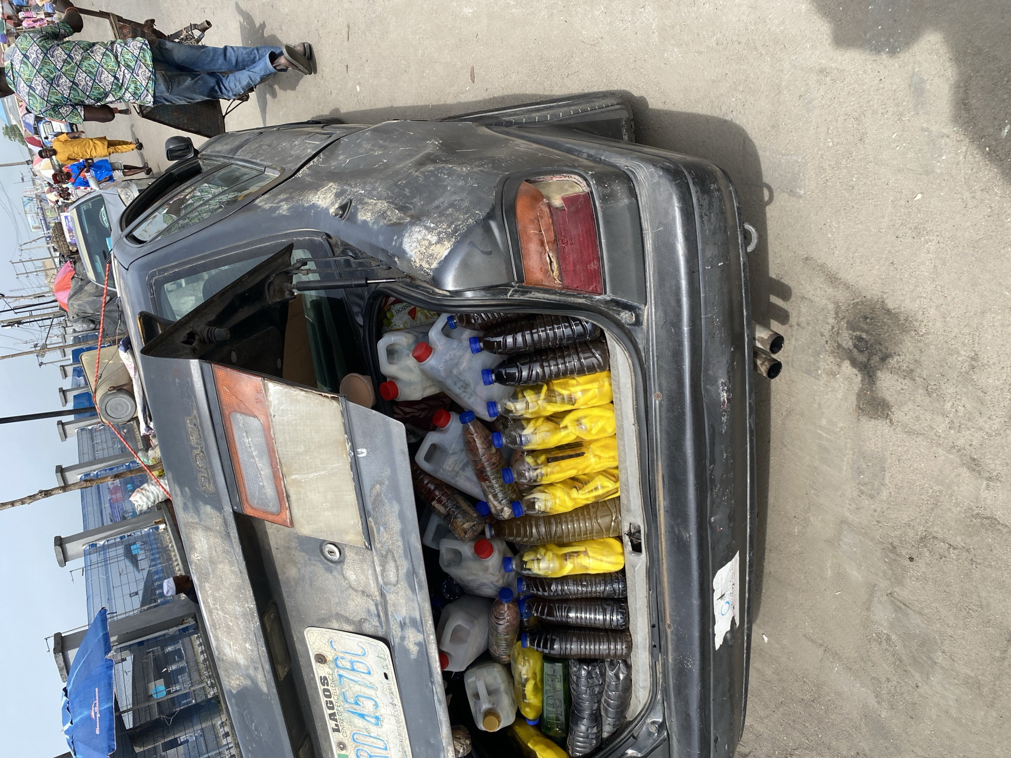 The image depicts the open trunk of a dark gray sedan car, possibly a Toyota, filled with numerous plastic bottles and jugs containing a mix of yellow and dark-colored liquids. The car's license plate is visible, indicating it is from Lagos, with the registration number RD 457 BC. The car appears to be in disrepair, with damage to the rear light and general wear and tear. The background shows an urban setting, likely a market or busy street scene, with people and other vehicles visible. The location is Lekki, Nigeria, as indicated by the license plate.