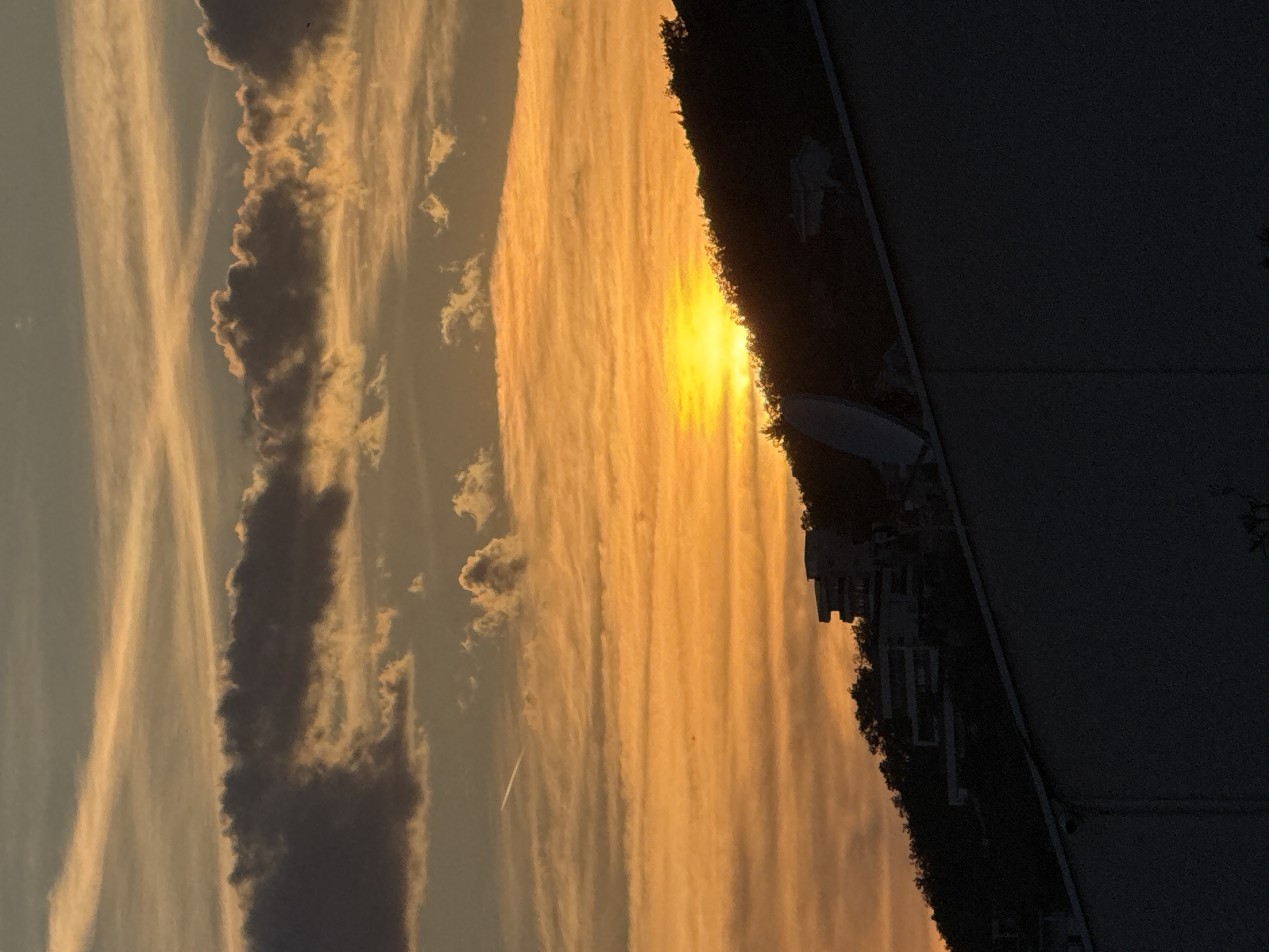 The image depicts a sunset viewed from Šušanj, Montenegro. The sky is filled with striated orange and yellow clouds, with some darker, less defined cloud formations on the left. The sun is positioned near the horizon, obscured by the clouds but emitting a bright glow. A contrail is visible in the sky. The silhouette of buildings and trees is seen along the horizon line, partially obscuring the sunset. A dark, undefined object occupies the top right portion of the frame.
