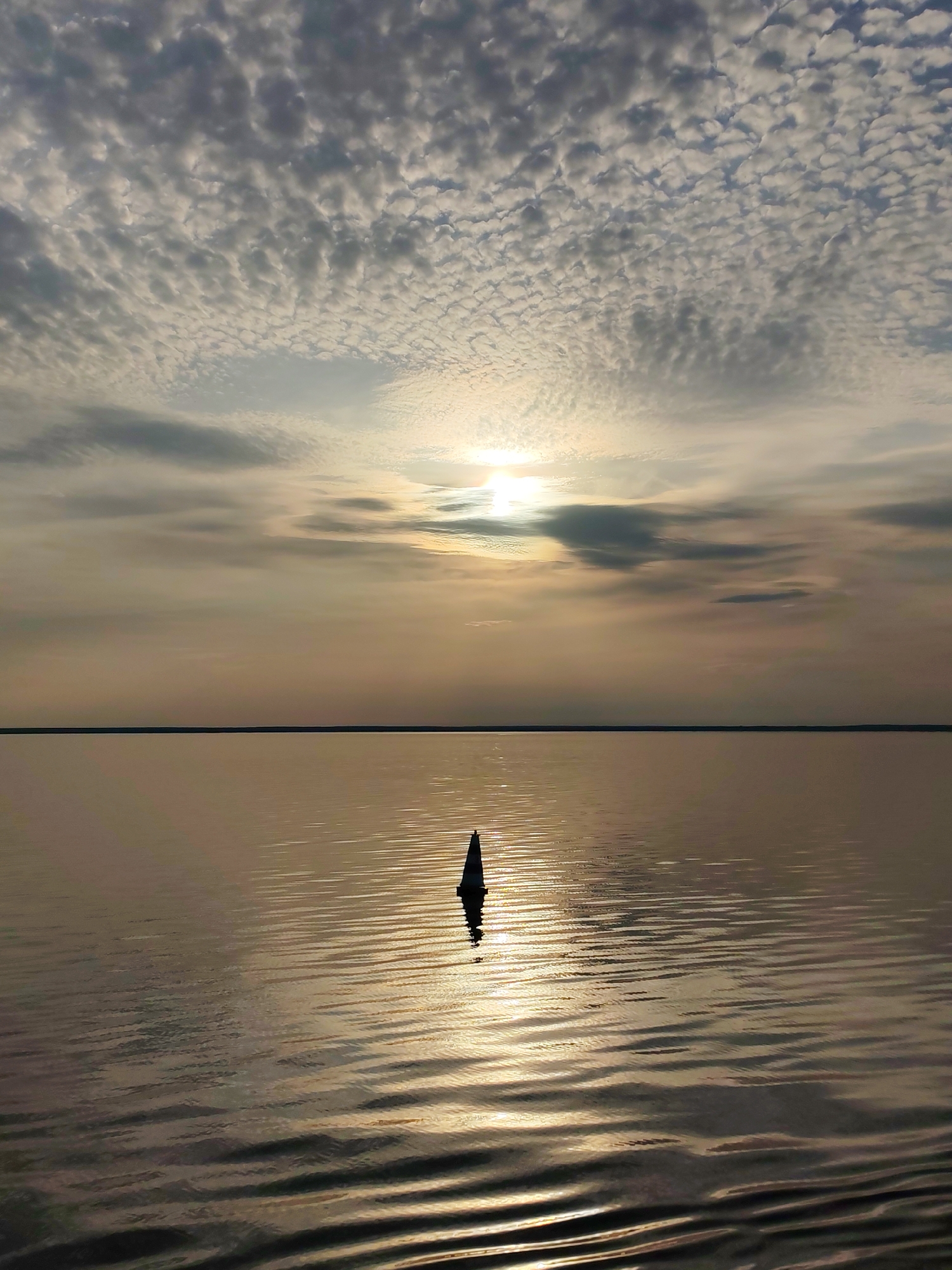 The image depicts a body of water under a cloudy sky, likely a lake or sea. A single, dark buoy or marker is visible in the middle ground, reflecting the light from the sky on the water's surface. The horizon line is distinct but obscured by atmospheric haze. The sun is partially obscured by clouds, creating a muted light. There is no evidence to confirm the location as Veregonets, Russia.
