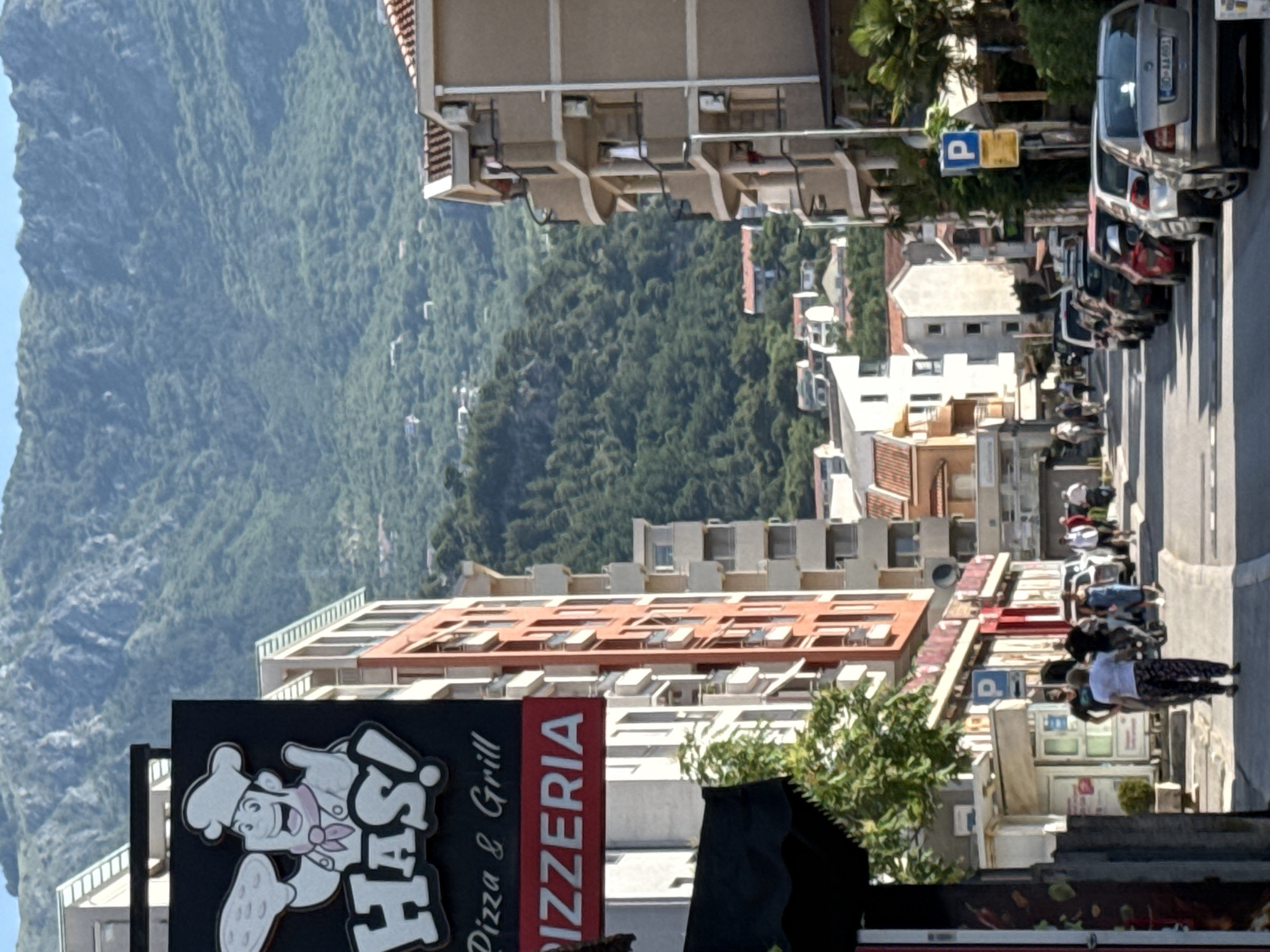 A vertical, eye-level shot captures a street scene in Budva, Montenegro, leading towards a lush, green mountain under a clear blue sky. On the left, a black sign with a cartoon chef holding a pizza advertises "HAS! Pizza & Grill" and "PIZZERIA" in bold white and red lettering, respectively. Adjacent to this, a tall, orange-and-white building with multiple balconies lines the street. Further down, a row of parked cars, including a silver Skoda, occupies the right side of the road. People are visible walking and interacting on the sidewalk and street. The architecture features multi-story buildings with balconies and tiled roofs, typical of coastal Montenegrin towns. The overall impression is a vibrant, sunlit street scene in a Mediterranean urban environment.