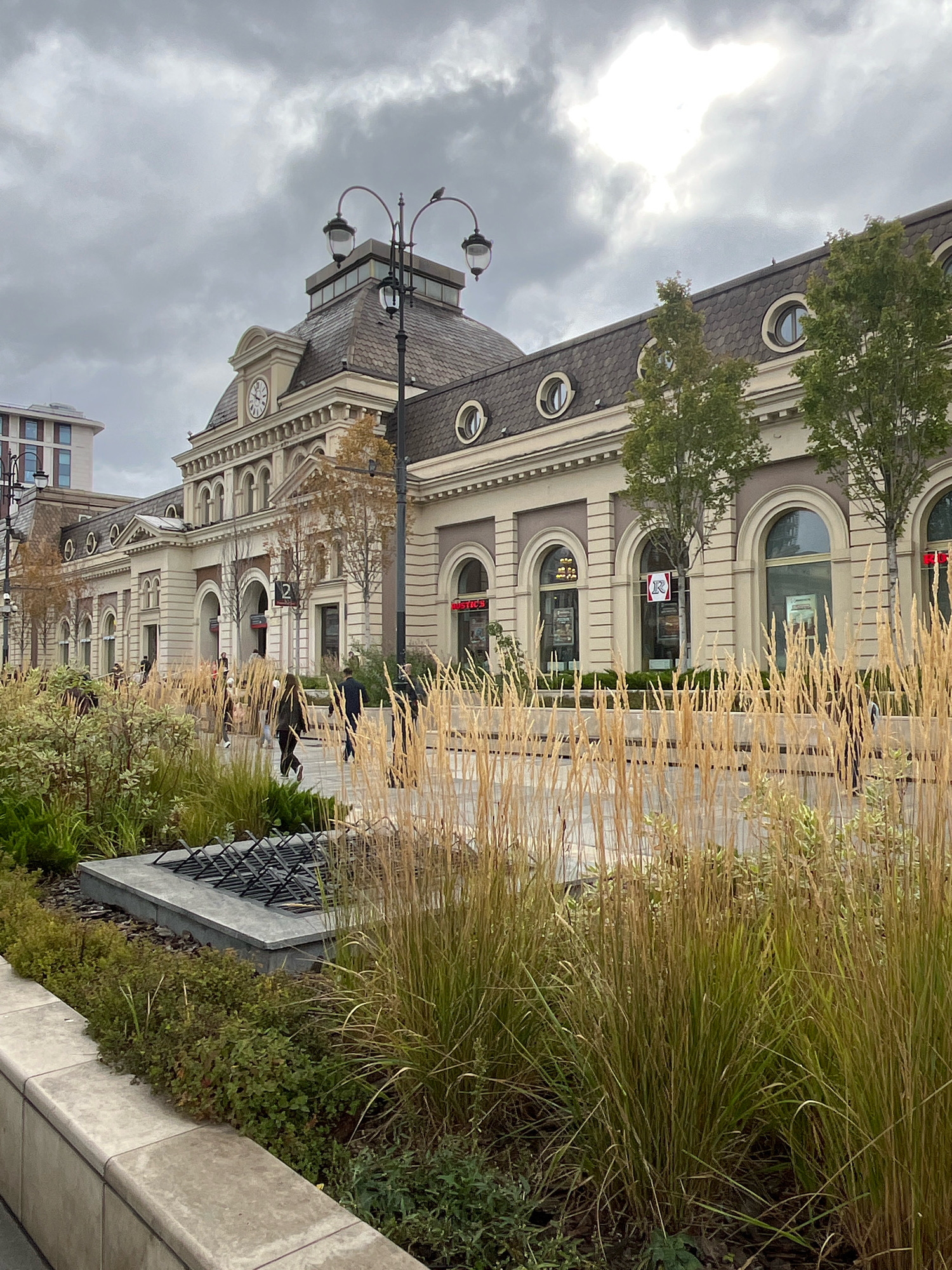 A public space in front of a building that appears to be a train station features a foreground of ornamental grass and low-lying bushes. In the mid-ground, several people are walking. The building itself is a light-colored structure with arched windows and a prominent clock on its facade. It is a cloudy day. The scene is located at Yaroslavsky Railway Station in Moscow, Russia.