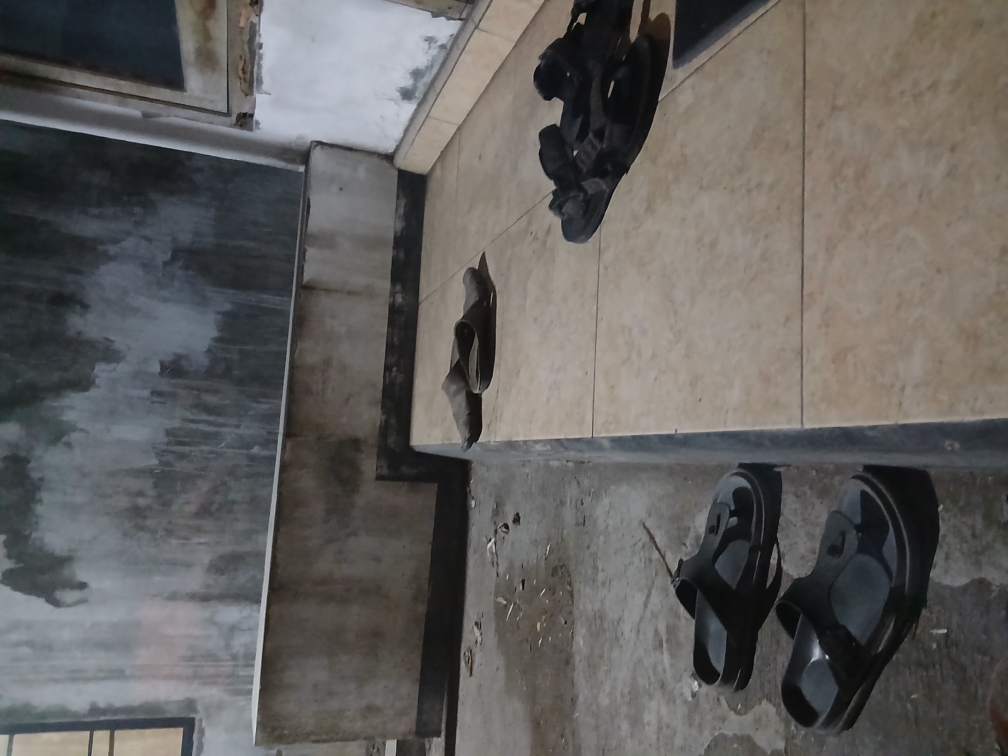 The image depicts an outdoor scene, possibly in Tenggarang, Indonesia, at what appears to be nighttime or dusk. The ground is a mix of concrete and tiled surfaces. A low concrete wall runs horizontally across the midground. Several pairs of sandals are scattered on the ground. In the foreground, two dark-colored sandals are placed on the concrete. Further back, on the tiled surface, a pair of dark sandals is visible, and another pair of lighter colored sandals is to their left. The background features a textured, weathered concrete wall with some dark stains, and a small window with a dark frame is visible on the left. To the right, a white pillar and a portion of another wall are present. The lighting is dim, casting shadows and creating a somber atmosphere.