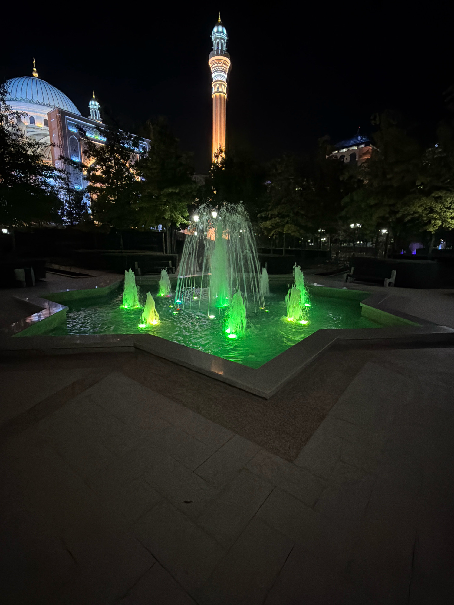 The image depicts a nighttime view of a fountain with green lights, located in the city of Shali, Russia. In the background, a minaret and a mosque are illuminated. The fountain is star-shaped and features multiple water jets. The surrounding area is paved with stone, and there are trees and benches visible. The lighting suggests a festive or decorative illumination of the area.