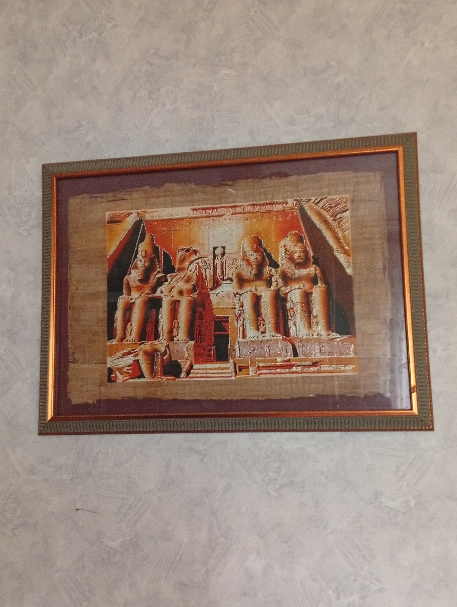 The image shows a framed artwork depicting ancient Egyptian statues on a textured wall. The artwork is a papyrus painting in shades of brown, red, and orange, featuring four seated figures in profile and other hieroglyphic elements. The frame is multi-layered, with an inner dark maroon mat and an outer textured metallic frame, possibly bronze or gold. The wall behind the artwork has a light gray, textured finish with a subtle pattern. The scene is indoors, and the lighting suggests a natural light source from the side. The photograph is taken at an angle, with the top of the frame slightly closer to the camera than the bottom. There are no people visible in the image. The location is identified as Pushkino, Russia.