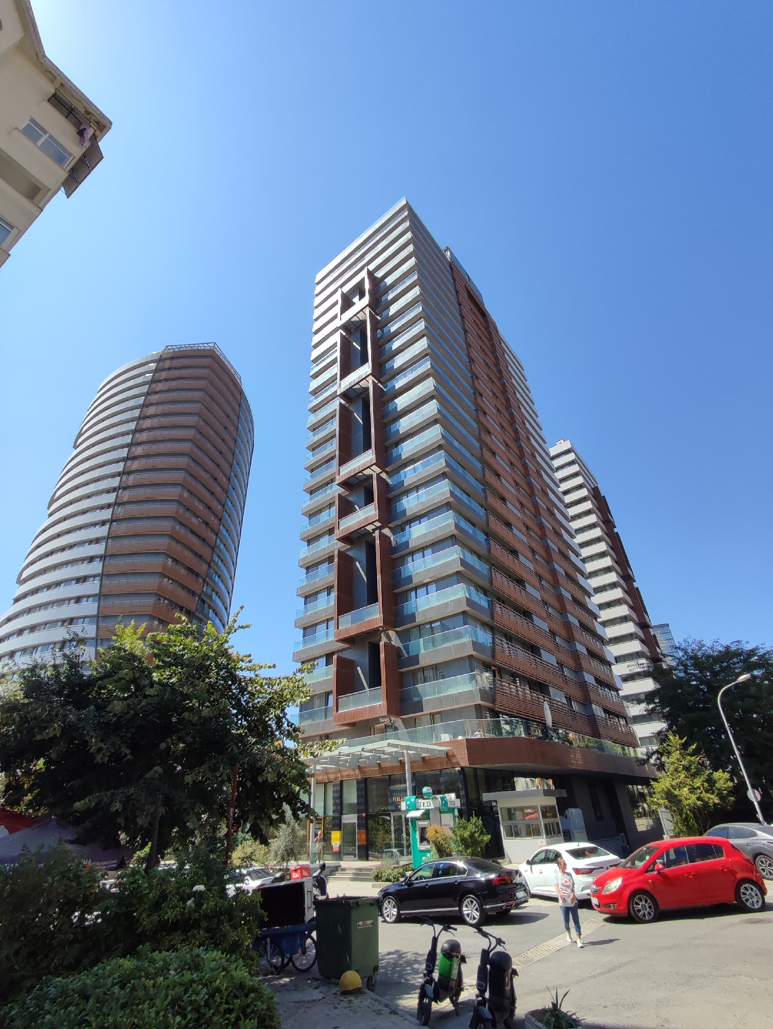 In Kadıköy, Turkey, a wide-angle photograph captures a bustling street scene dominated by modern architecture under a clear blue sky. Two prominent, tall buildings with distinct designs stand out: a curved, light-colored skyscraper on the left, and a more angular, brown-and-glass tower on the right, featuring stacked rectangular cutouts.
The street level is active with traffic and pedestrian activity. A bright red hatchback is parked on the right side of the road, alongside a white sedan and a black sedan. A young woman in a striped shirt and jeans is walking in the street between the red and white cars. Two electric scooters are parked on the sidewalk on the left. In the foreground, lush green trees and bushes partially obscure a blue cart with a black container and a green trash bin. The facades of the buildings feature glass balconies and decorative paneling. Businesses with storefronts are visible at the base of the taller buildings, including a branch of "TEB" bank with an ATM. A streetlight extends into the frame from the right. The overall impression is one of a vibrant urban environment.