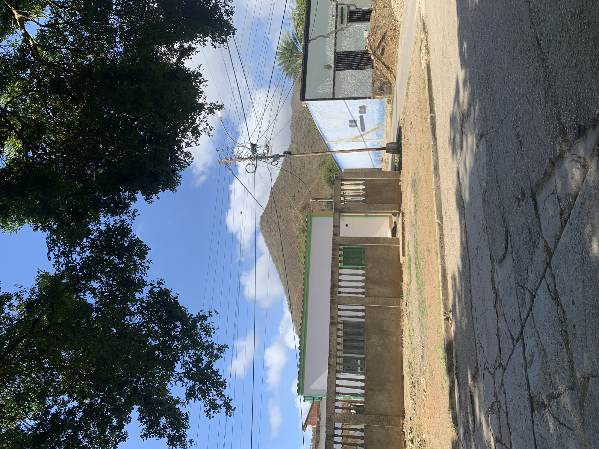 In Santa Ana, Venezuela, a street scene unfolds under a bright, cloudy sky, framed by the dark, leafy branches of trees overhead. In the foreground, a cracked asphalt road reflects dappled shadows. Beyond the road, a low, rough concrete wall runs horizontally across the frame, topped by a series of decorative balusters. Behind this wall, a white building with a green trim is visible, its windows partially obscured. To the right, a light blue building with dark barred windows and stone accents stands, a pile of dirt and gravel adjacent to it. A utility pole with numerous crisscrossing wires and a street lamp stands near the blue building. In the background, a large, dry, mountainous landscape rises under the sky.