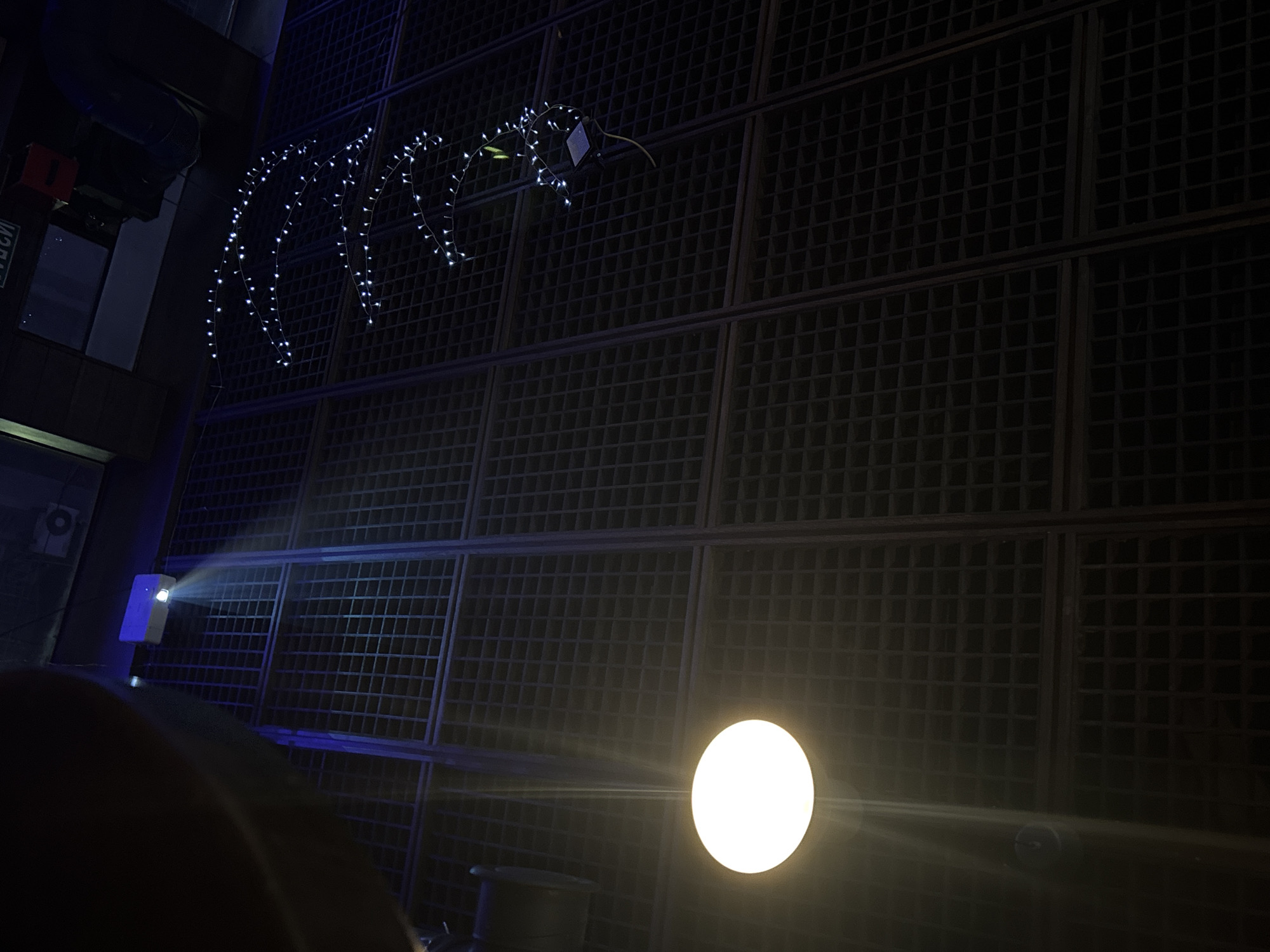 A low-angle indoor photograph shows a room with a dark gridded wall, a string of lights, and a bright light source.
The dark, gridded wall occupies the right and upper portions of the frame. The grid appears to be made of dark metal. A string of lights is hung near the top left of the grid.
To the left of the gridded wall is a dark corner, where a white projector and a dark object are located.
A bright, white, spherical light source is in the lower center of the frame, emitting rays of light.
The image appears to have been taken at night. The location is not explicitly stated but could be in Belgrade, Serbia.