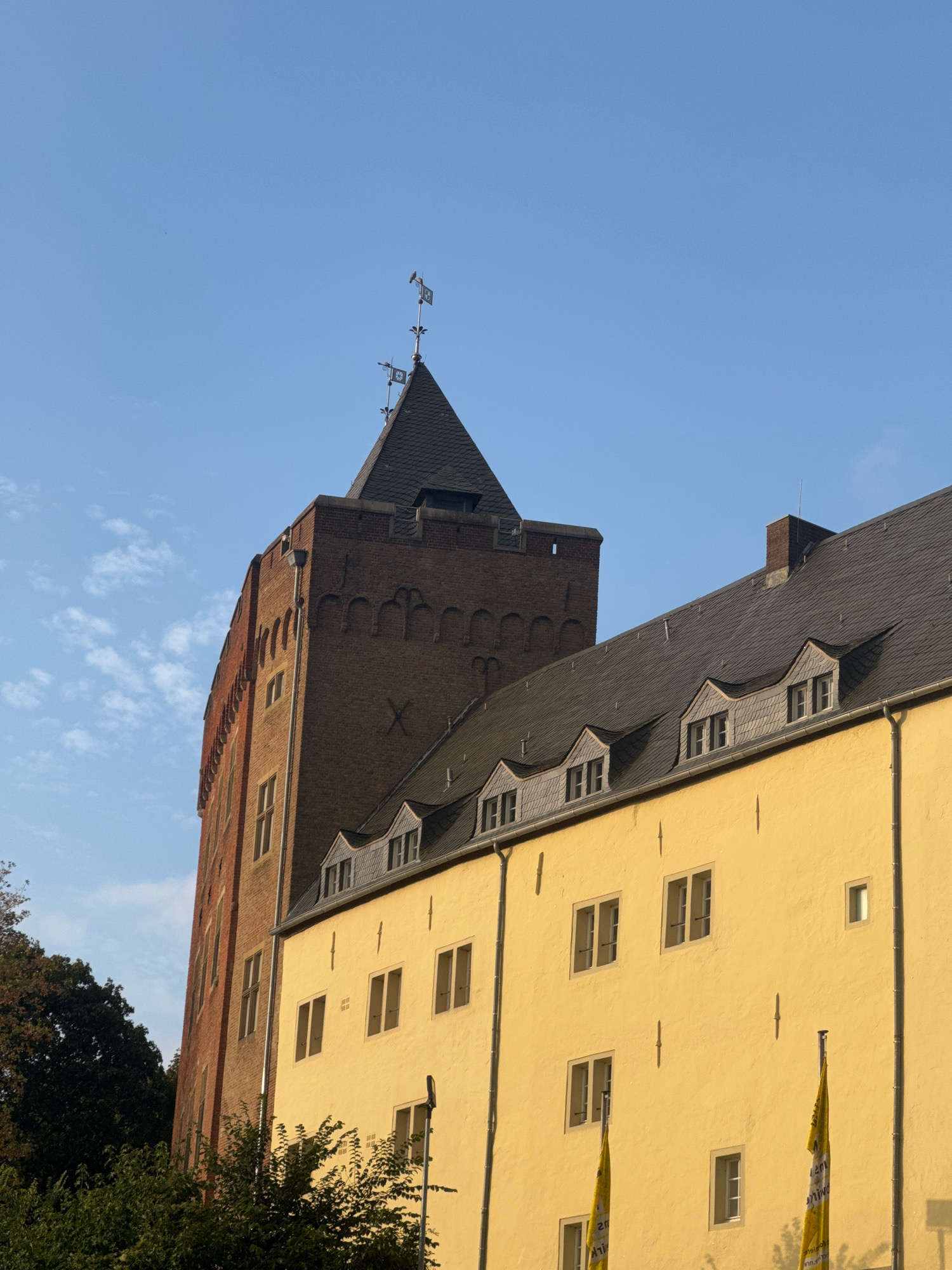 The image shows a close-up view of the exterior of the Cleves Castle (Schloss Cleves) in Cleves, Germany, under a clear blue sky with a few wispy clouds. The castle features a tall, reddish-brown brick tower with arched decorative elements and a dark, sloped roof topped with a weather vane. Adjacent to the tower is a yellow building with a dark slate roof, characterized by multiple dormer windows with slate roofing and casement windows. The yellow facade has evenly spaced, rectangular windows and some vertical decorative elements. Two yellow flags with text, partially visible, are planted in front of the yellow building. Dense green trees are visible on the left side of the image, partially obscuring the lower portion of the brick tower. The lighting suggests it is daytime, with sunlight casting shadows and highlighting the textures of the buildings.