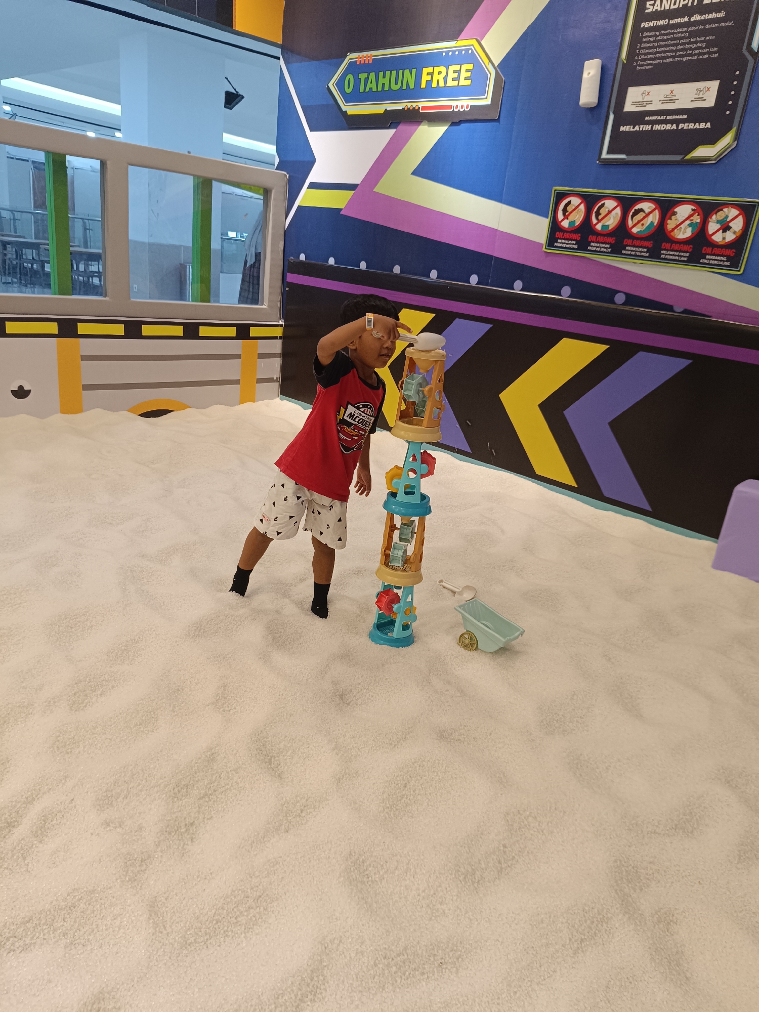 A young boy in a red shirt and patterned shorts stands in a shallow sandpit, his right hand raised to his forehead as if shielding his eyes. He is facing away from the viewer, towards a colorful toy tower structure with various gears and buckets. To his right, a small blue toy wheelbarrow and a shovel rest on the sand. The sandpit is vast and appears to be indoors, with a wall decorated with a large blue and purple chevron pattern behind the boy. A sign with Indonesian text and graphics is affixed to the wall. The sandpit is likely part of a children's play area in a mall or entertainment center in Sorong, Indonesia.