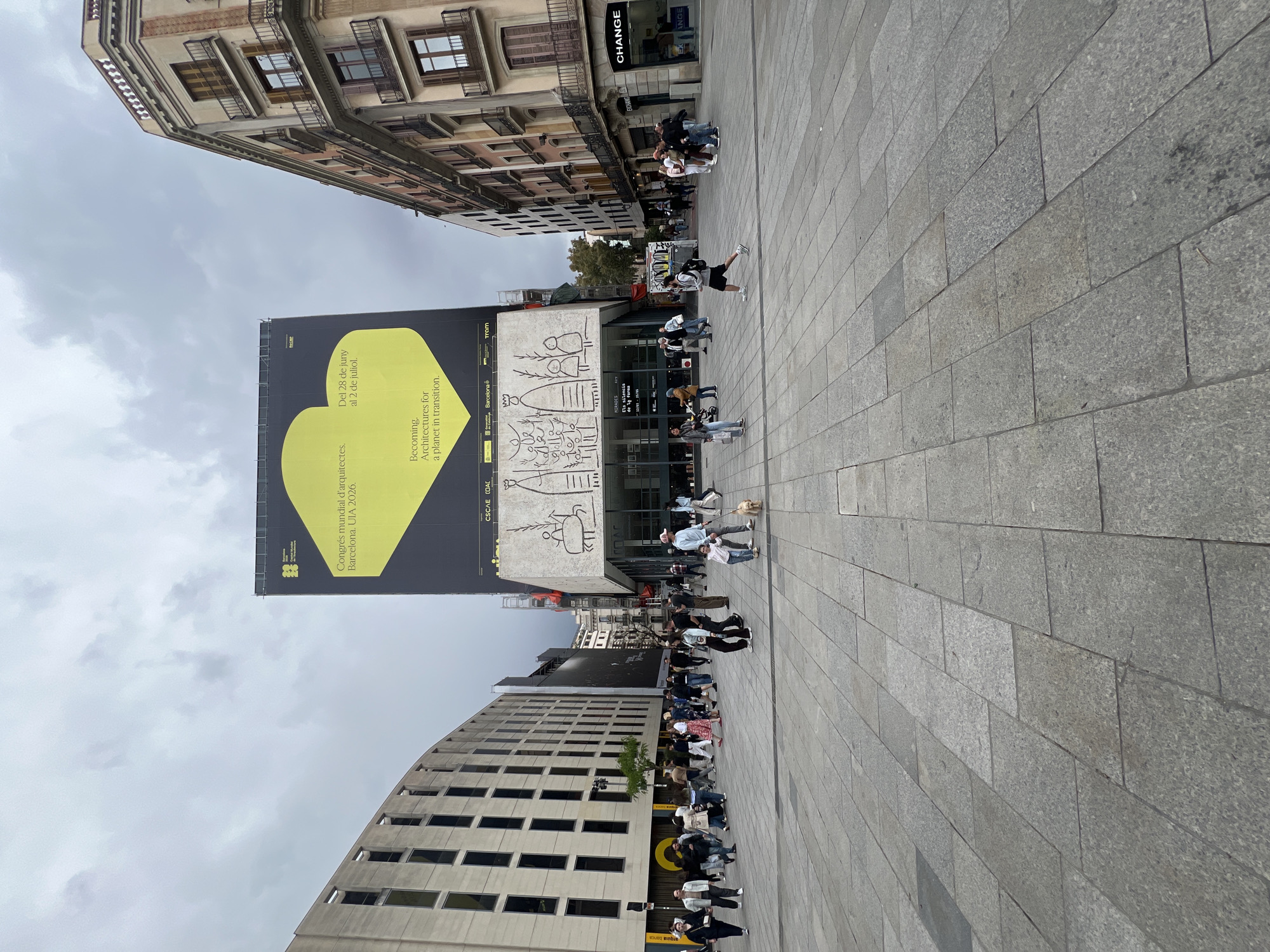 The image shows a wide pedestrian plaza in Barcelona, Spain. The ground is paved with large, rectangular grey stones. A large building on the left has a curved facade and numerous windows, and it has a sign for "arquia banca." In the middle is a taller building with a large yellow-and-black banner promoting the "Congrés mundial d'arquitectes. Barcelona. UIA 2026." It states the event will be held from June 28th to July 2nd with the theme "Becoming. Architectures for a planet in transition." The building also has a facade with drawings on it. On the right is a more traditional building with a "CHANGE" store. Many people are present, walking and standing around the plaza. The sky is overcast.