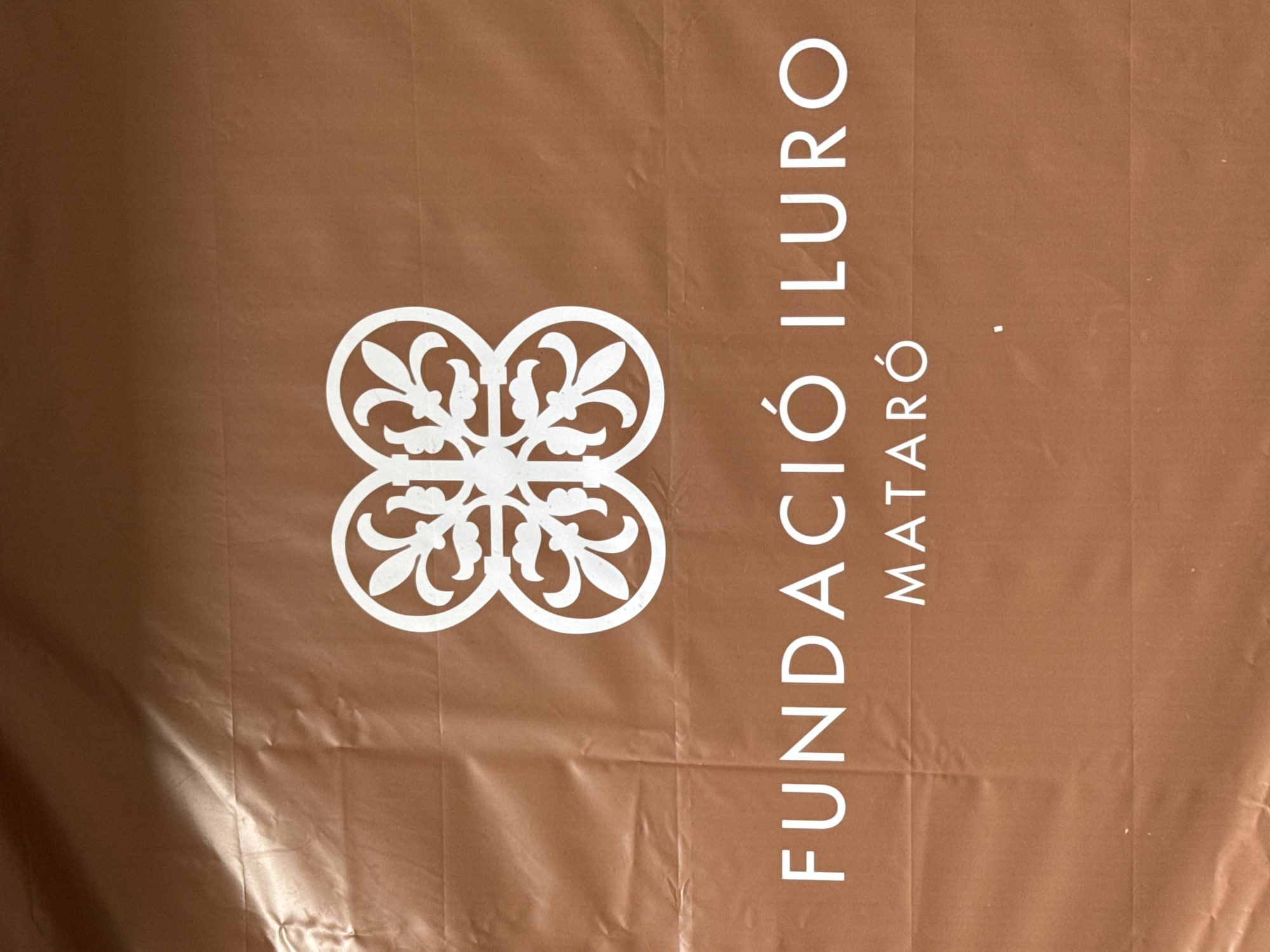 The image shows a white logo on a brown, wrinkled surface. The logo consists of a stylized floral or quatrefoil design at the top, and text below. The text reads "FUNDACIÓ ILURO" and, centered below that, "MATARÓ". The surface appears to be a banner or sign. The location is confirmed by the text "MATARÓ", which is the name of a city in Catalonia, Spain.