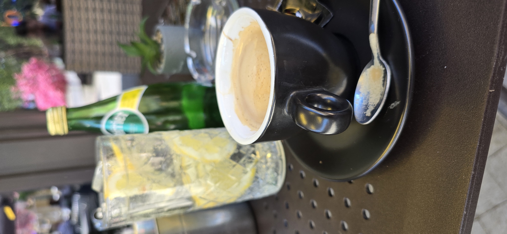 A close-up, medium-shot, rotated eye-level outdoor shot shows a black cup of coffee in a black saucer with a silver spoon on a brown, perforated surface. Behind the coffee is a glass of water with lemon slices and a green glass bottle. The background is blurred, showing some greenery and pink flowers. The setting appears to be a cafe or outdoor seating area. The city is Sofia, Bulgaria.