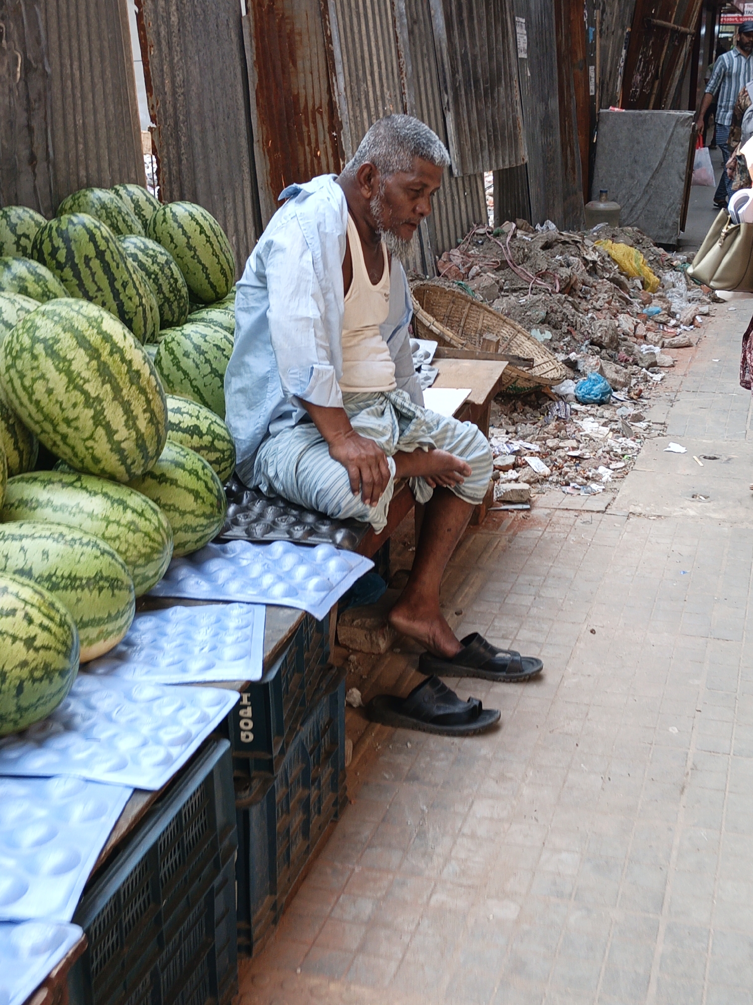 A man is sitting on a bench in the city of Dhaka, Bangladesh, next to a pile of watermelons. He is wearing a white tank top, a light blue shirt, and a striped wrap. The bench is made of wooden planks and is placed on top of blue trays in black crates. The watermelons are stacked next to him. Behind the man is a corrugated metal wall. To the right of the bench, there is a pile of debris, including plastic bags, broken bricks, and other refuse. The ground is paved with light-colored tiles. A person wearing a striped shirt is walking away from the scene in the background.