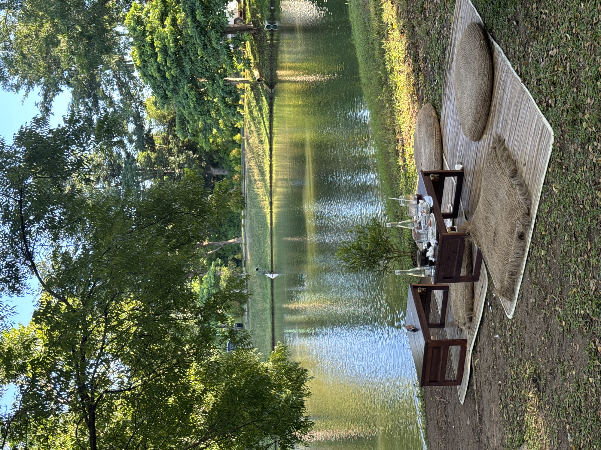 A picnic is set up beside a canal or narrow lake in Kuta Selatan, Indonesia. A woven mat serves as the base, with round pillows and a fringed throw. A small table with two stools holds glassware, plates, and bottles. Trees and foliage surround the water.