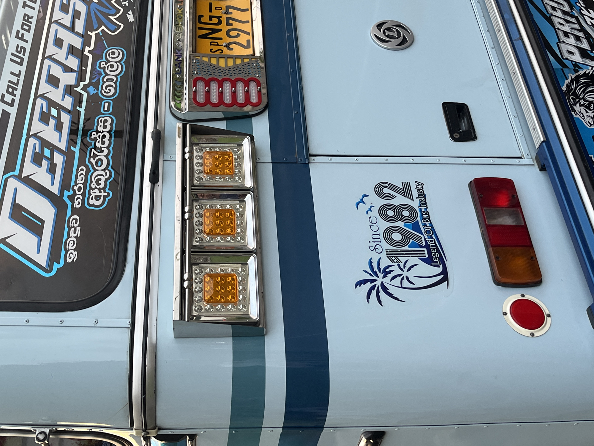 The image shows a close-up of the side of a vehicle, likely a bus, in the city of Galle, Sri Lanka. The vehicle is light blue with darker blue stripes. The vehicle has a sign written in English and Sinhalese with the name "JEERAS" and the route "Akurassa-Galle". There are multiple lights, including rectangular orange turn signals, red brake lights, and a license plate that reads "SPNG 2977". A logo with the text "Since 1982 Legend Of Bus Industry" and a palm tree design is also visible.