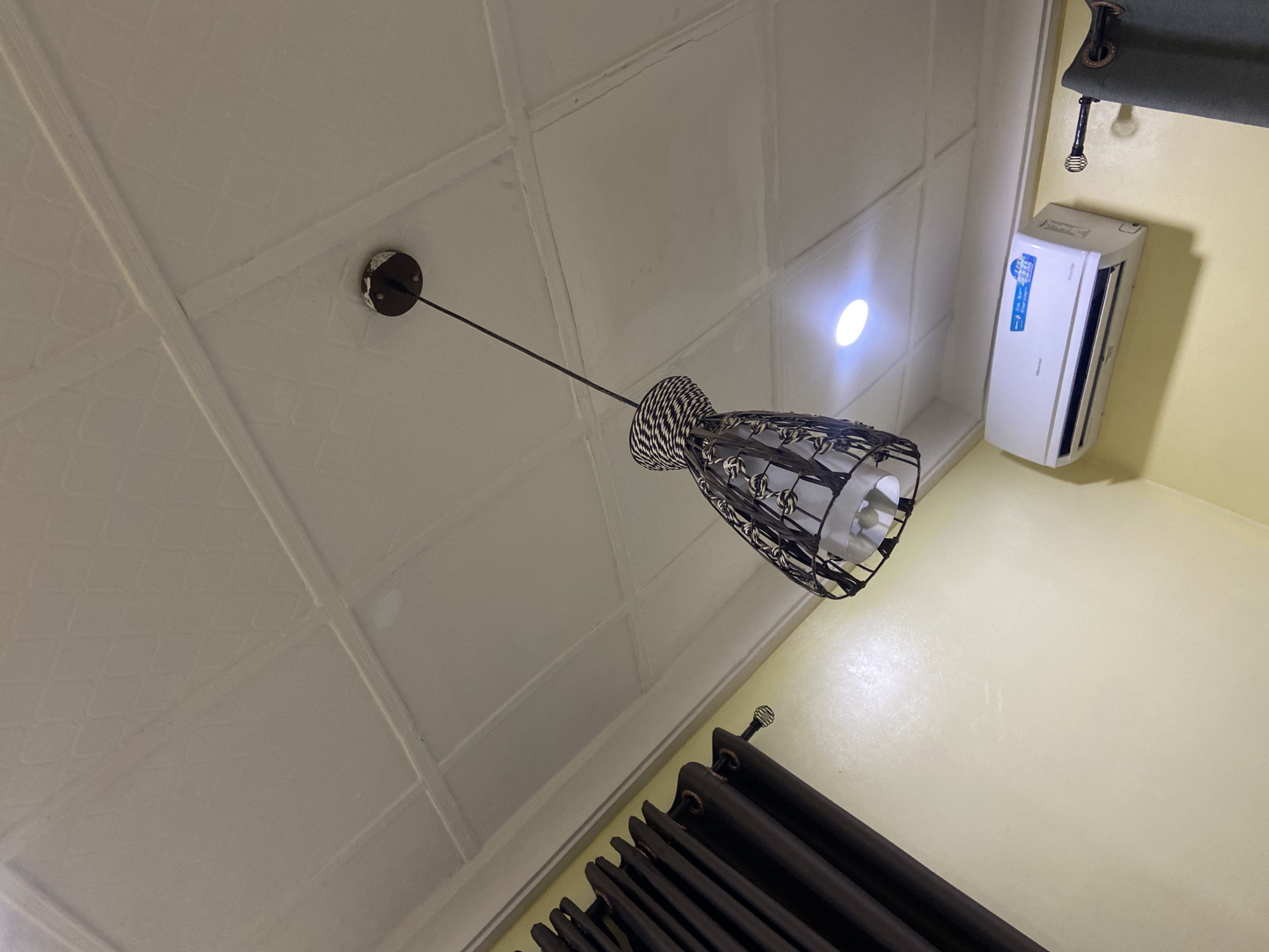 The image shows an indoor scene, likely a room, with a focus on the ceiling and upper walls. A decorative ceiling light fixture with a black metal cage and a woven top hangs from the ceiling. The ceiling itself is white and appears to be made of square panels with raised edges. On the wall below the ceiling, a portion of a dark brown curtain is visible on the left side of the image. To the right, a white air conditioning unit is mounted on a pale yellow wall. A light from the air conditioner's display is visible. A curtain rod with decorative finials is also present on the right. The location is Gura Top, Nigeria.