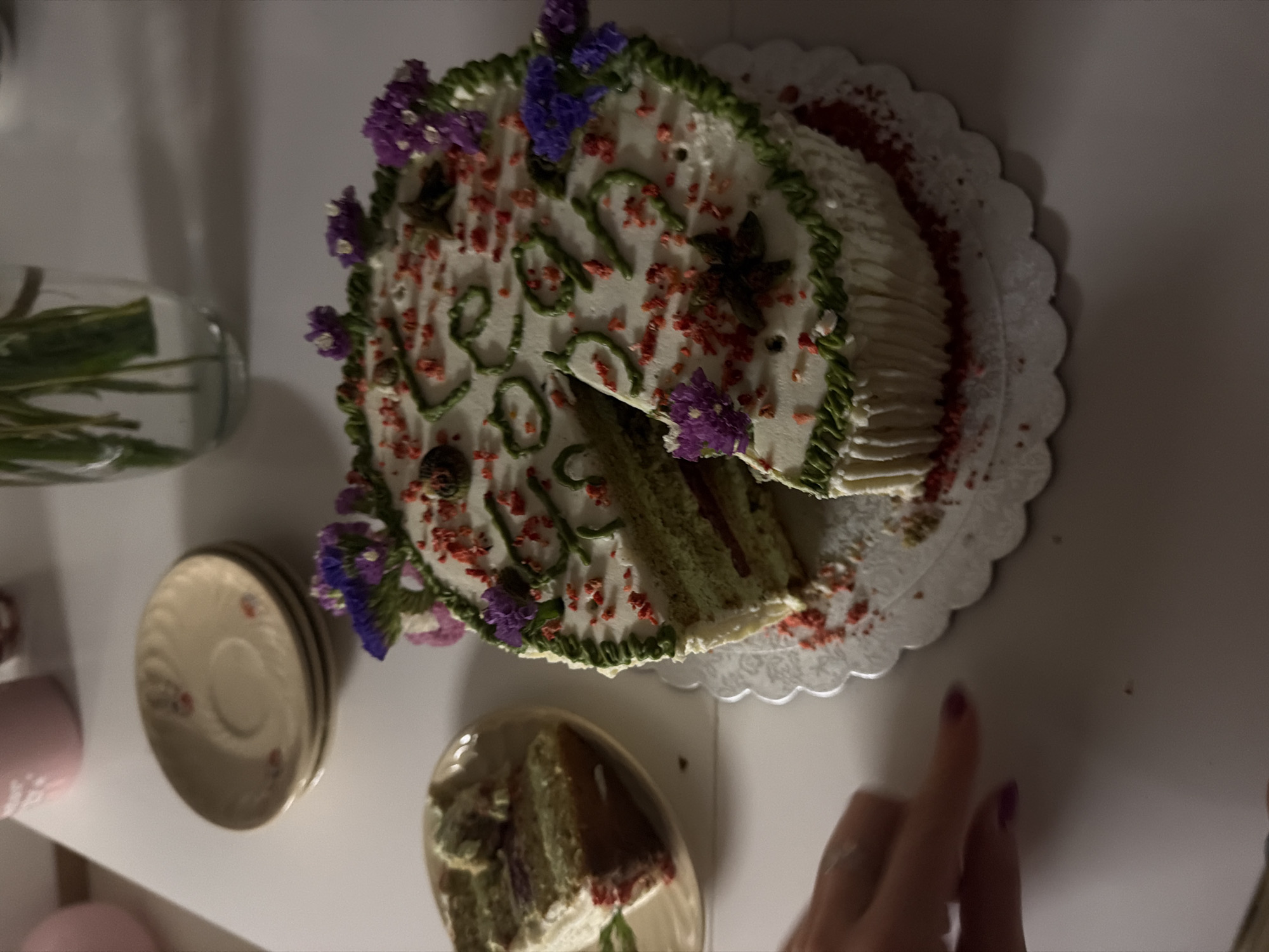 The image shows a partially eaten cake on a white table, with a slice already removed and placed on a small plate to the left. The cake is decorated with white frosting, green icing swirls, purple flowers, and red sprinkles. A hand with purple nail polish is visible reaching towards the cake. In the background, there are stacked saucers and a vase with greenery. The scene appears to be a domestic setting, possibly a home or a cafe, located in Vracar Urban Municipality, Serbia.