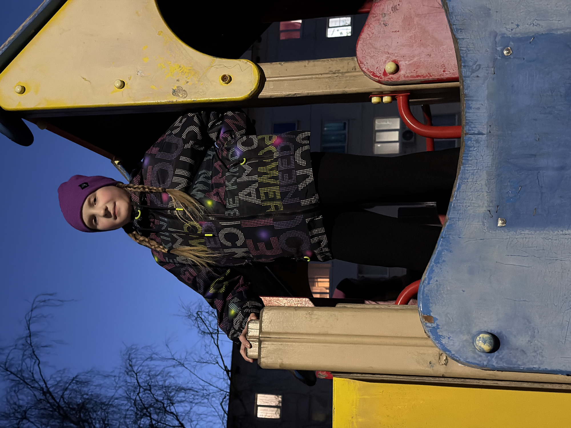 A young girl with blonde braided hair and a purple beanie is playing on playground equipment. She is wearing a black jacket with colorful patterns and black pants. She is holding onto a white ledge of the playground structure. The playground equipment is made of metal and painted in various colors, including yellow, red, and blue. Behind her, there is a building with illuminated windows, suggesting it is nighttime. Bare tree branches are visible in the lower left corner. The image was taken in Chaikovsky, Russia.