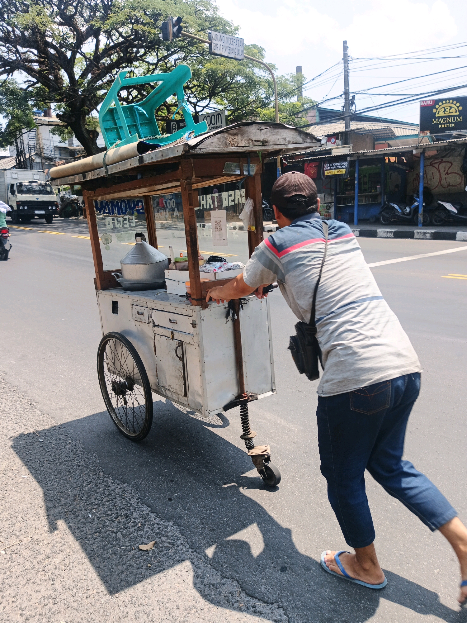 A man wearing a t-shirt, jeans, a cap, and flip-flops pushes a food cart down a street in Bandung City, Indonesia. The food cart is made of metal and wood, with a large pot and other cooking utensils visible inside. The man's right hand is on the cart, and his body is angled away from the camera as he walks. A large tree with intricate branches shades part of the street, and a traffic light is visible in the background. The street is paved, with yellow lane markings. Other vehicles and buildings are present in the distance, suggesting an urban environment. The image is captured on a bright, sunny day, casting a distinct shadow from the food cart and the man. The man is walking on the right side of the road.