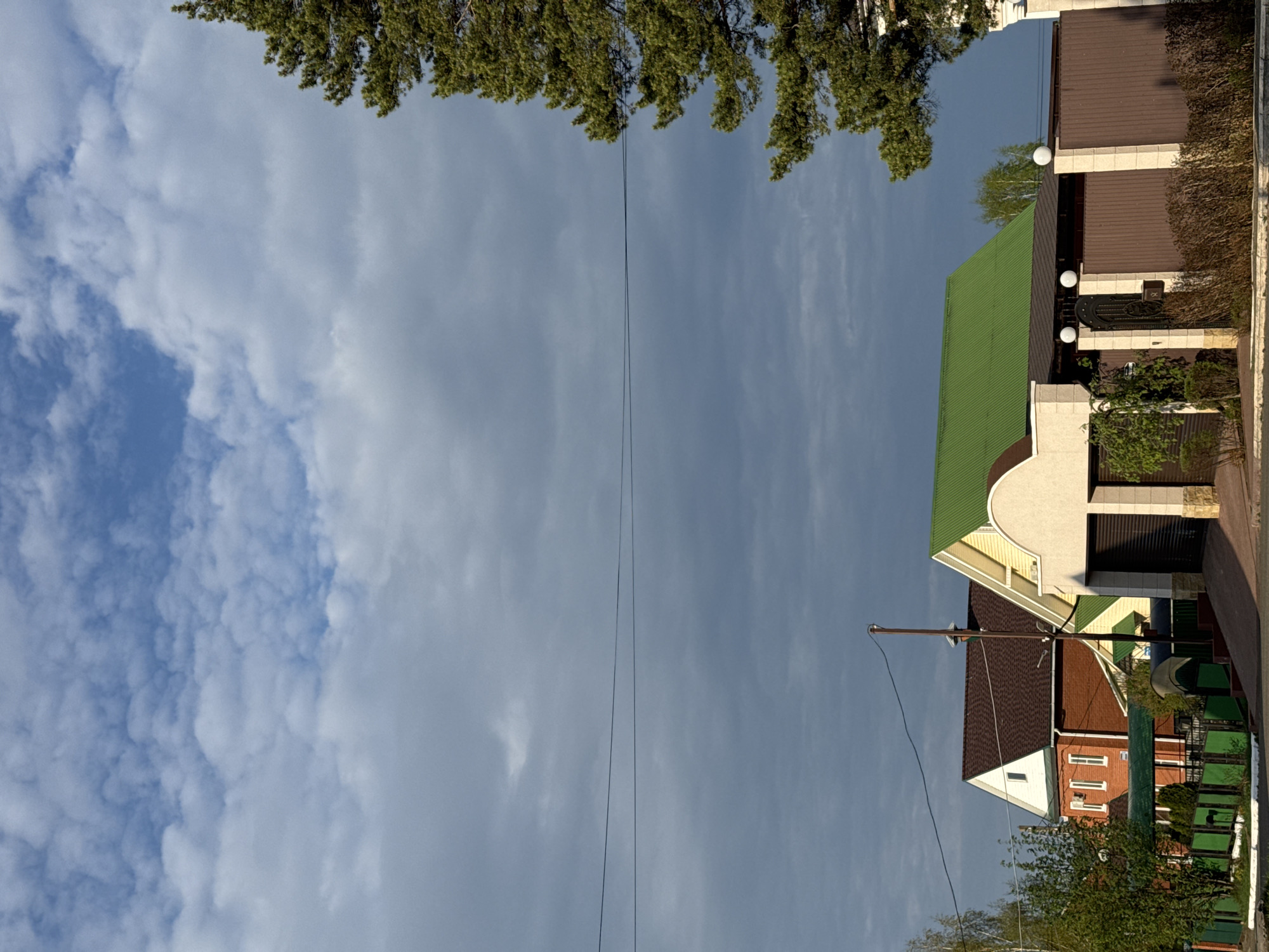 The image shows a street scene in Прикамский, Russia. The sky is partially cloudy with varying shades of blue and white. At the top of the frame, there's a line of green tree foliage. On the right side of the image, there are several buildings with different roof colors including green, brown, and red. There's also a utility pole with wires running across the sky. The architecture appears to be residential. The image is taken from a low angle, looking upward.