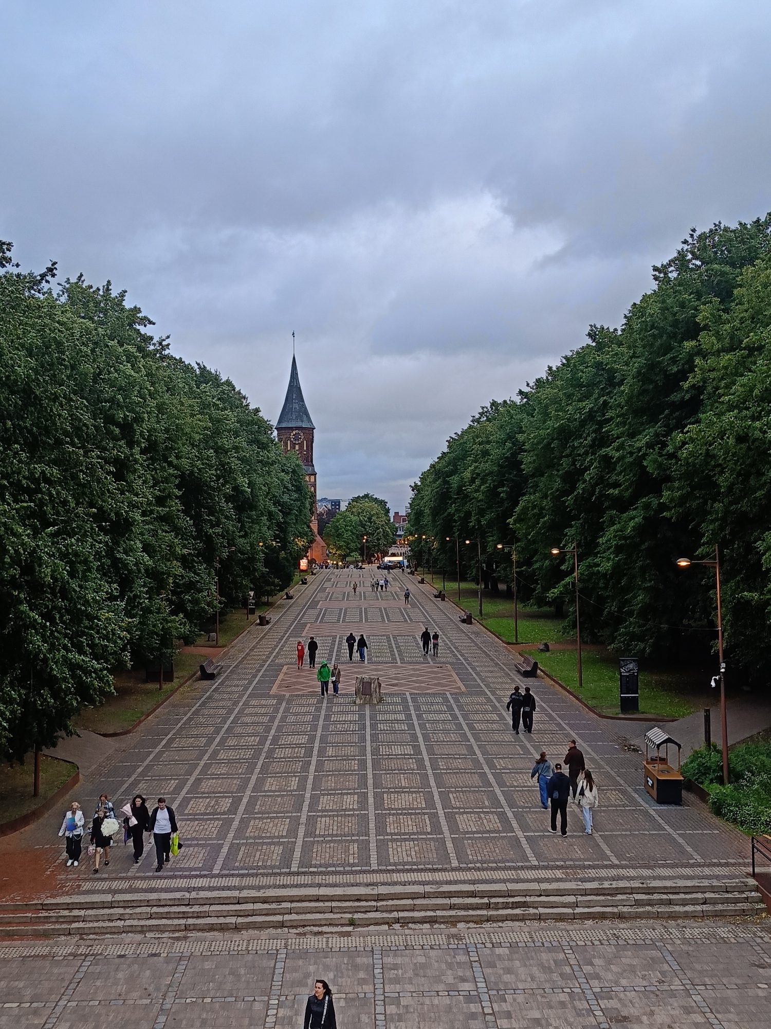 The image depicts a wide, paved promenade lined with mature trees on either side, leading towards a prominent church with a tall, spired bell tower. This location is identified as Kant Island in Kaliningrad, Russia, with the church being the Cathedral of Christ the Saviour. The sky is overcast, suggesting a cloudy day. Several individuals are walking along the promenade and on the paved steps leading up to it. On the promenade, people are seen in small groups and individually, some carrying bags. The paving stones are laid in a patterned grid. Lampposts are visible along the sides of the promenade, and benches are placed between the trees. In the background, beyond the church, other buildings and trees can be seen.