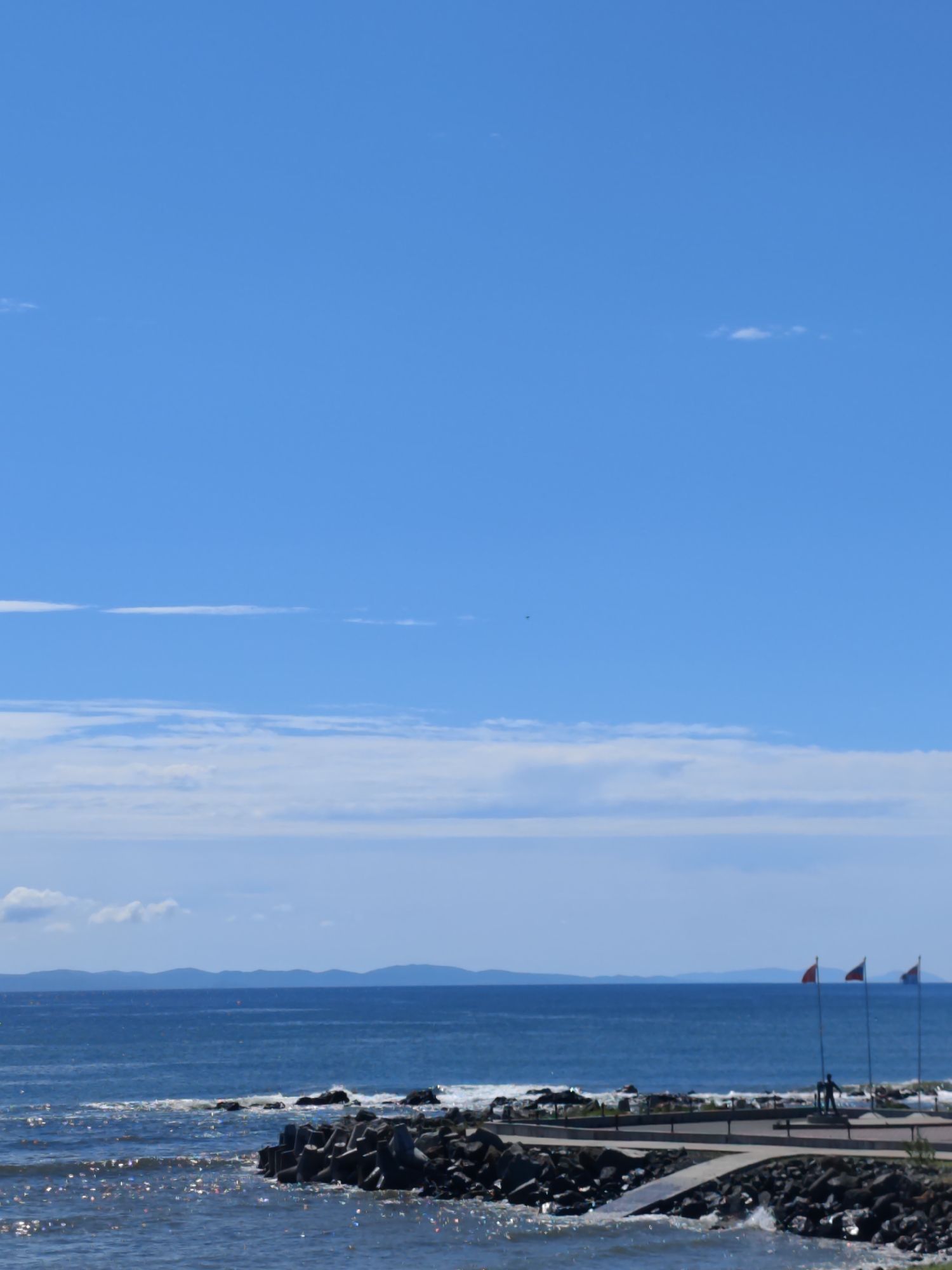 The image depicts a coastal scene with a clear blue sky and a calm ocean. In the foreground, a rocky breakwater is visible, with concrete tetrapods forming a barrier against the waves. The water is a deep blue and appears slightly choppy. In the mid-ground, a paved walkway runs along the edge of the breakwater. Three flagpoles stand on the walkway, each with a flag. The flags are red, white, and blue, suggesting a national or regional affiliation. Two silhouetted figures are standing on the walkway near the flagpoles, appearing to be looking out at the ocean. In the background, a distant landmass with rolling hills is visible, shrouded in a light haze. The overall impression is one of a serene and picturesque coastal landscape. The location appears to be a publicly accessible area, possibly a park or promenade, designed to offer scenic views of the sea.