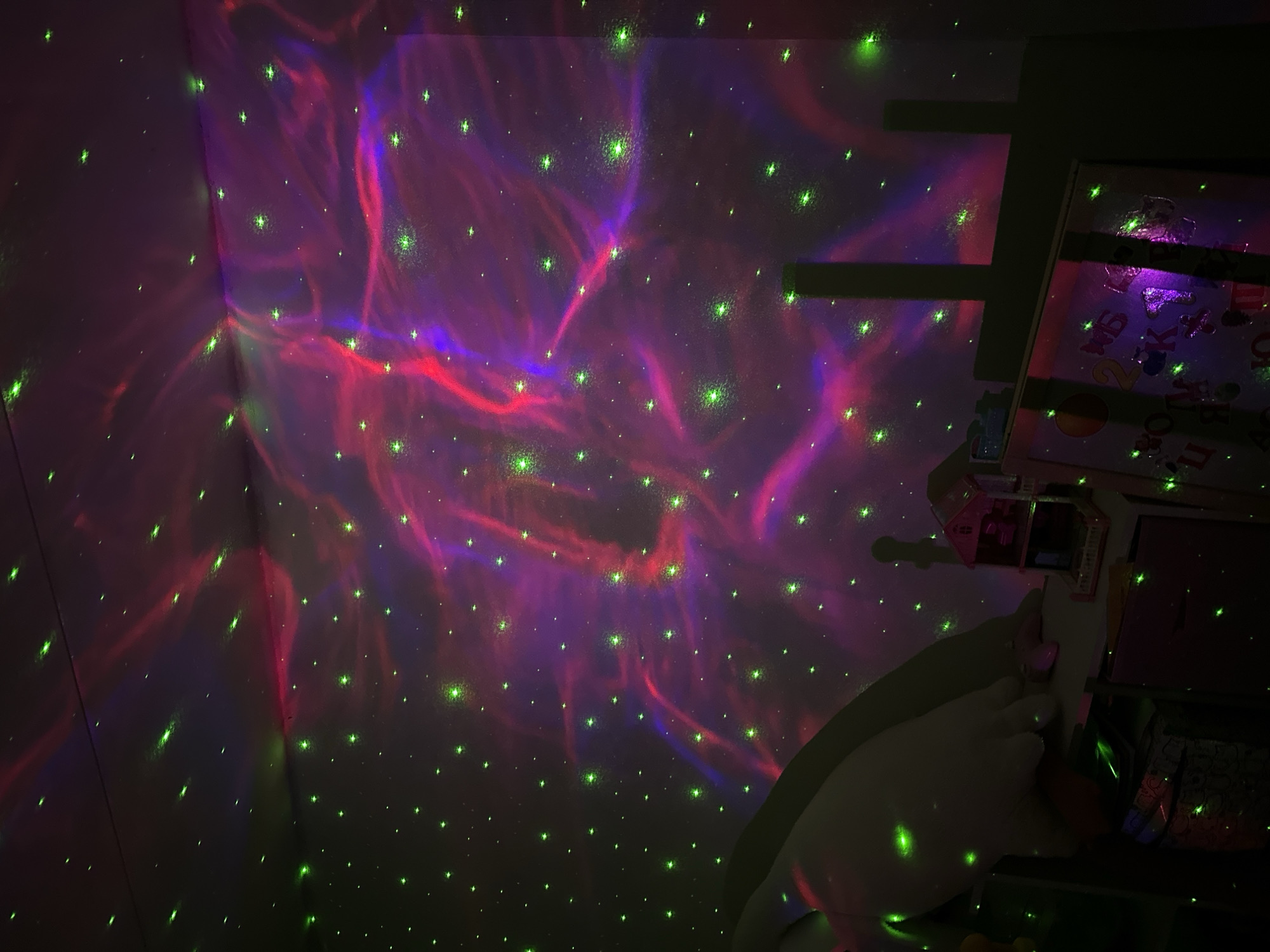 The image shows an interior corner of a room illuminated by a star projector. The walls and ceiling are covered in red and purple nebula patterns with numerous green star-like projections. To the right, there is a partial view of a shelf or piece of furniture, appearing to contain items like toys. No people are visible. No information suggests the location is in the city of Chaikovsky, Russia.