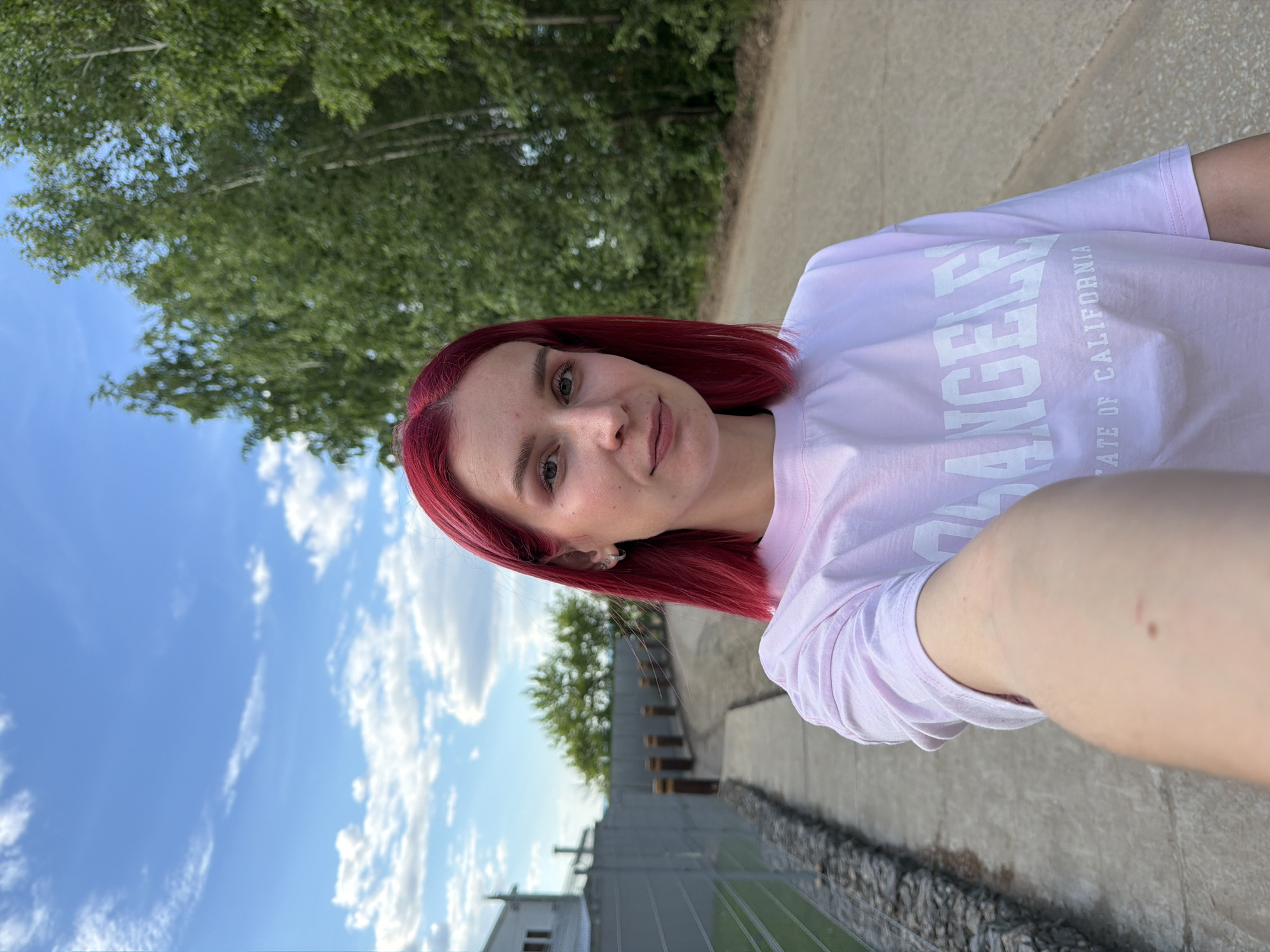 A young adult female, appearing in her 20s-30s, is depicted in a selfie taken outdoors in Chaikovsky, Russia. She has bright red, straight, shoulder-length hair and fair skin. Her eyes are dark, and her eyebrows are light-colored. She wears a light pink, short-sleeved t-shirt with white block letters on the front, partially visible as "ANGELES" and "STATE OF CALIFORNIA." Small silver hoop earrings with a small charm are visible on her right earlobe. Her right arm is extended into the foreground, holding the camera, and displays a small dark mole and two faint red marks on the inner forearm.
The scene is set on a clear day with a blue sky and scattered white cumulus clouds. To the subject's left, a gray metal fence runs parallel to a paved path. Behind the fence, a stone-filled gabion structure is visible at ground level. Further back, a light-colored building with dark windows is partially visible, followed by green foliage. Several short, brown cylindrical bollards or posts are spaced along the paved path. To the subject's right, a dirt or gravel road/path runs parallel to the paved path, flanked by a dense stand of green deciduous trees, some of which appear to be birches. The overall environment suggests a park, rural road, or a suburban area with significant green space.