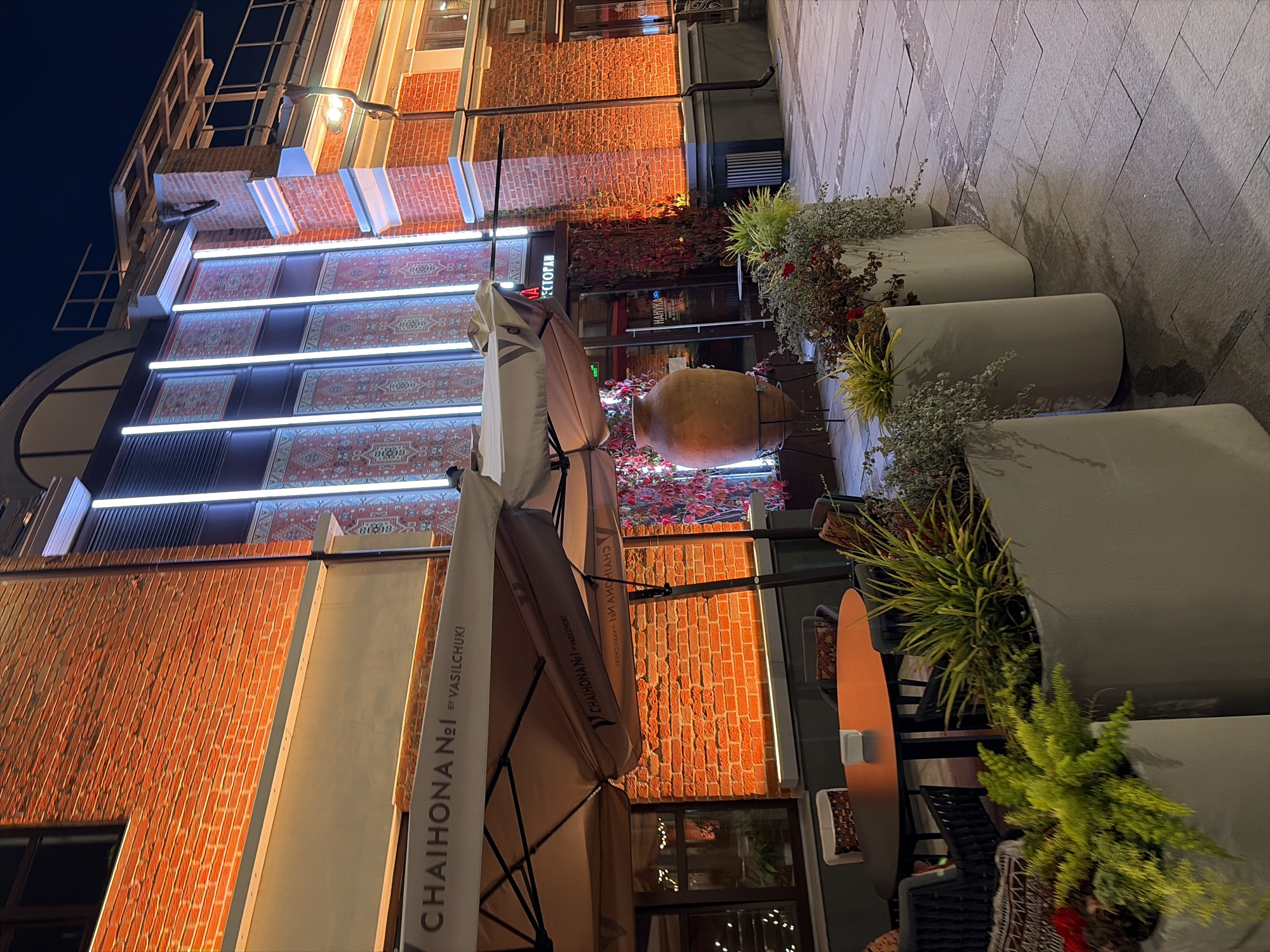 The image depicts the exterior of a restaurant at night. The building facade is constructed of red brick and features large vertical windows illuminated with white light, interspersed with panels decorated with intricate red and blue patterns that resemble Persian rugs. The restaurant's name, "CHAIHONA №1 BY VASILCHUKI," is visible on a white awning stretched over an outdoor seating area.
The outdoor seating area includes a round orange table with chairs and planters filled with greenery. A large, terracotta-colored amphora stands near the entrance. The ground is paved with stone tiles. The overall ambiance suggests a vibrant urban setting, likely a street-level establishment in a city. Based on the restaurant chain name, this is likely a location in Moscow, Russia. No people are visible in the image.