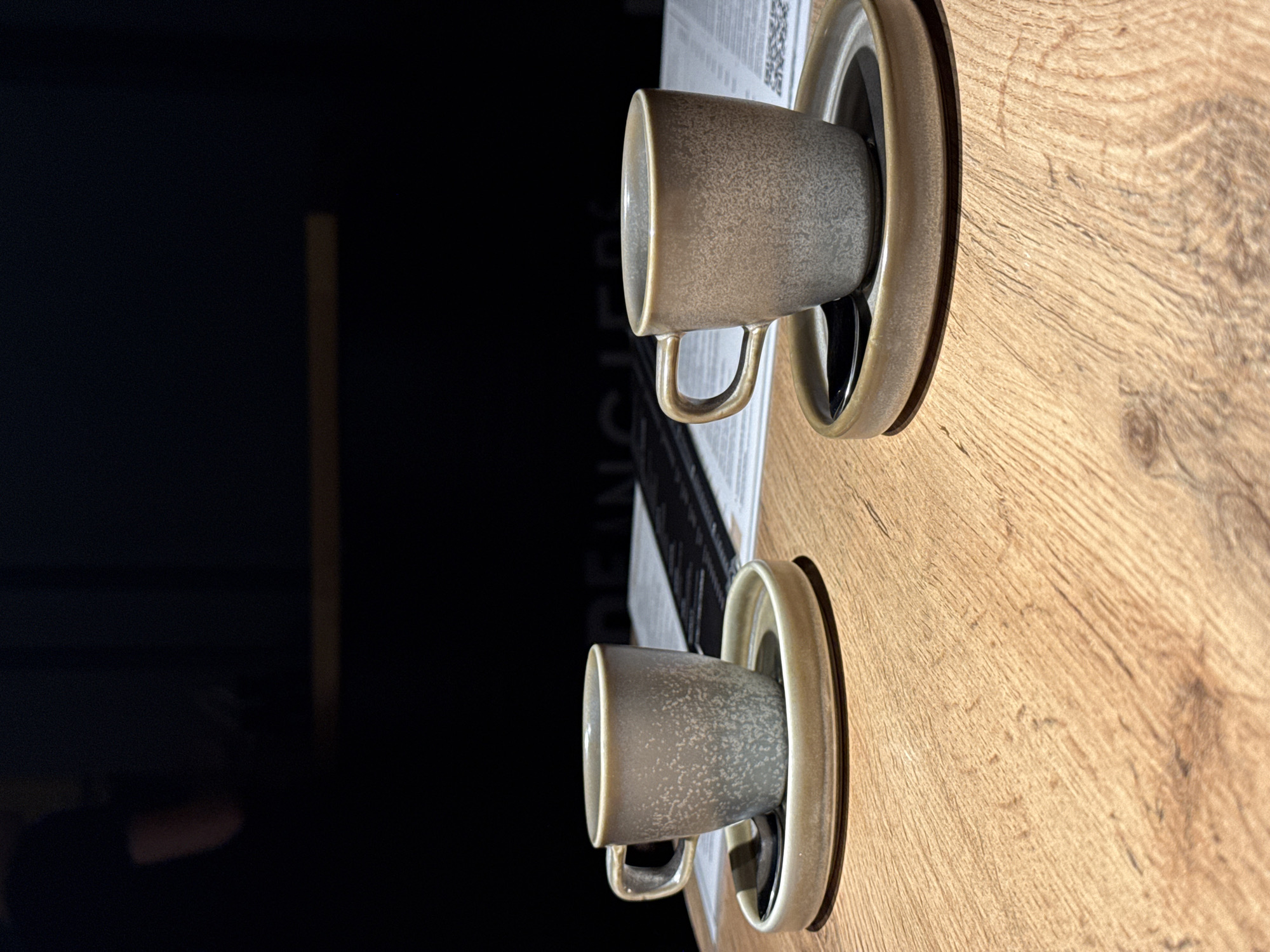 The image shows two light grey speckled ceramic cups with handles sitting on matching saucers. They are placed on a light brown wooden table, next to a menu. The location is most likely a cafe or restaurant in the city of Chaikovsky, Russia.