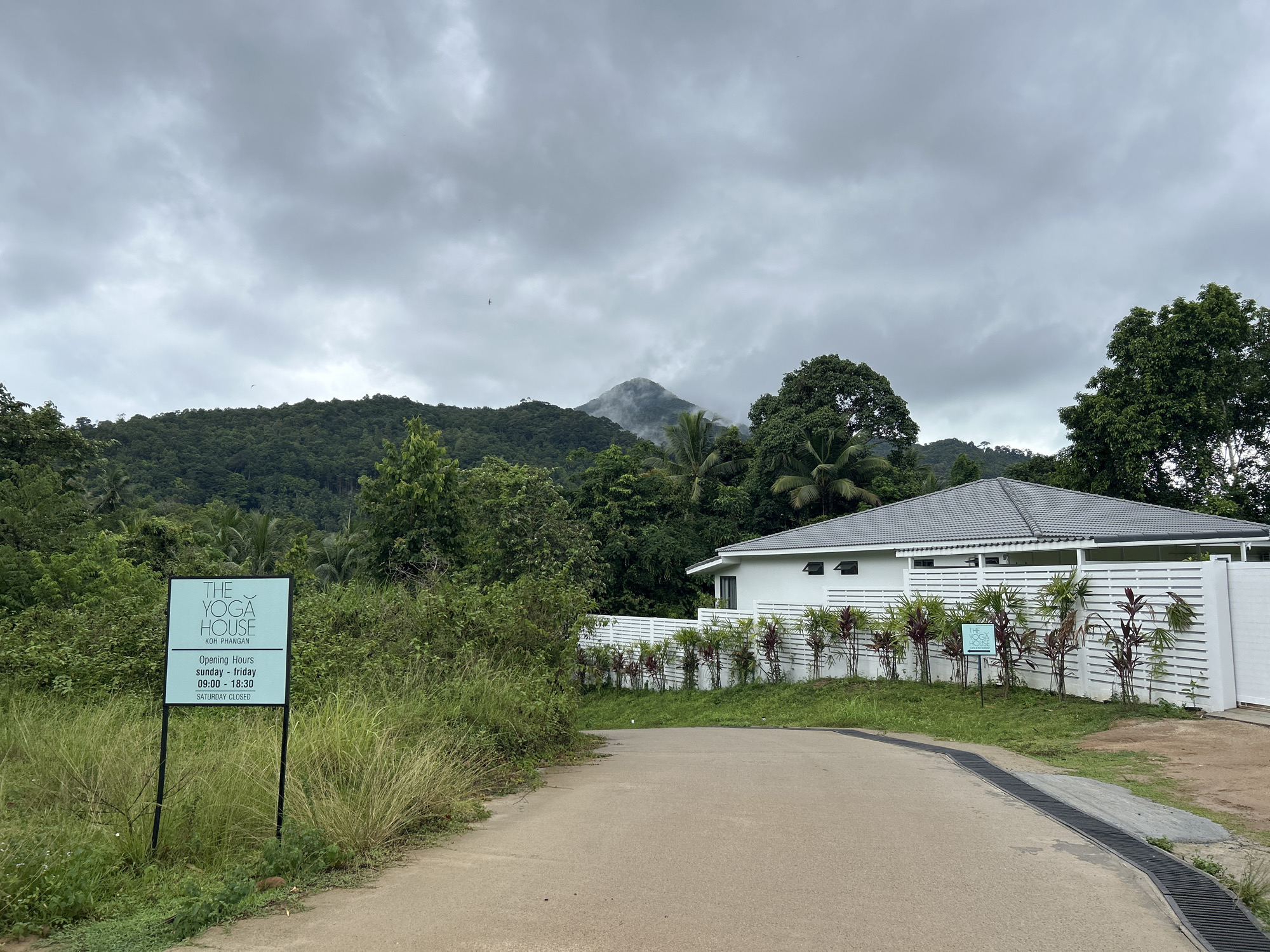 This media file captures a serene, tropical landscape dominated by lush greenery and a modern white building under a heavily overcast sky. In the foreground, a paved road or driveway curves to the right, bordered by wild tall grass and smaller shrubs on the left.
To the left of the road, a prominent sign stands, displaying "THE YOGA HOUSE KOH PHANGAN" in a clean, modern font. Below this, the opening hours are listed: "sunday - friday 09:00 - 18:30" and "SATURDAY CLOSED".
The midground features a stark white building with a grey tiled roof, possibly "The Yoga House" itself, partially obscured by dense vegetation. A white slatted fence runs along the front of the building, decorated with rows of red and green tropical plants. A smaller blue sign with "THE YOGA HOUSE" and an arrow pointing right is visible near the fence.
In the background, a majestic, forested mountain or large hill rises, its peak shrouded in mist or low clouds, indicating high humidity or recent rain. The sky above is entirely covered in thick, grey clouds, suggesting an overcast day. No direct sunlight is visible, giving the scene a soft, diffused light. The overall atmosphere is tranquil and conducive to relaxation or wellness activities. The dense tropical foliage reinforces the setting in Koh Phangan, Thailand. No people are visible in the image.
