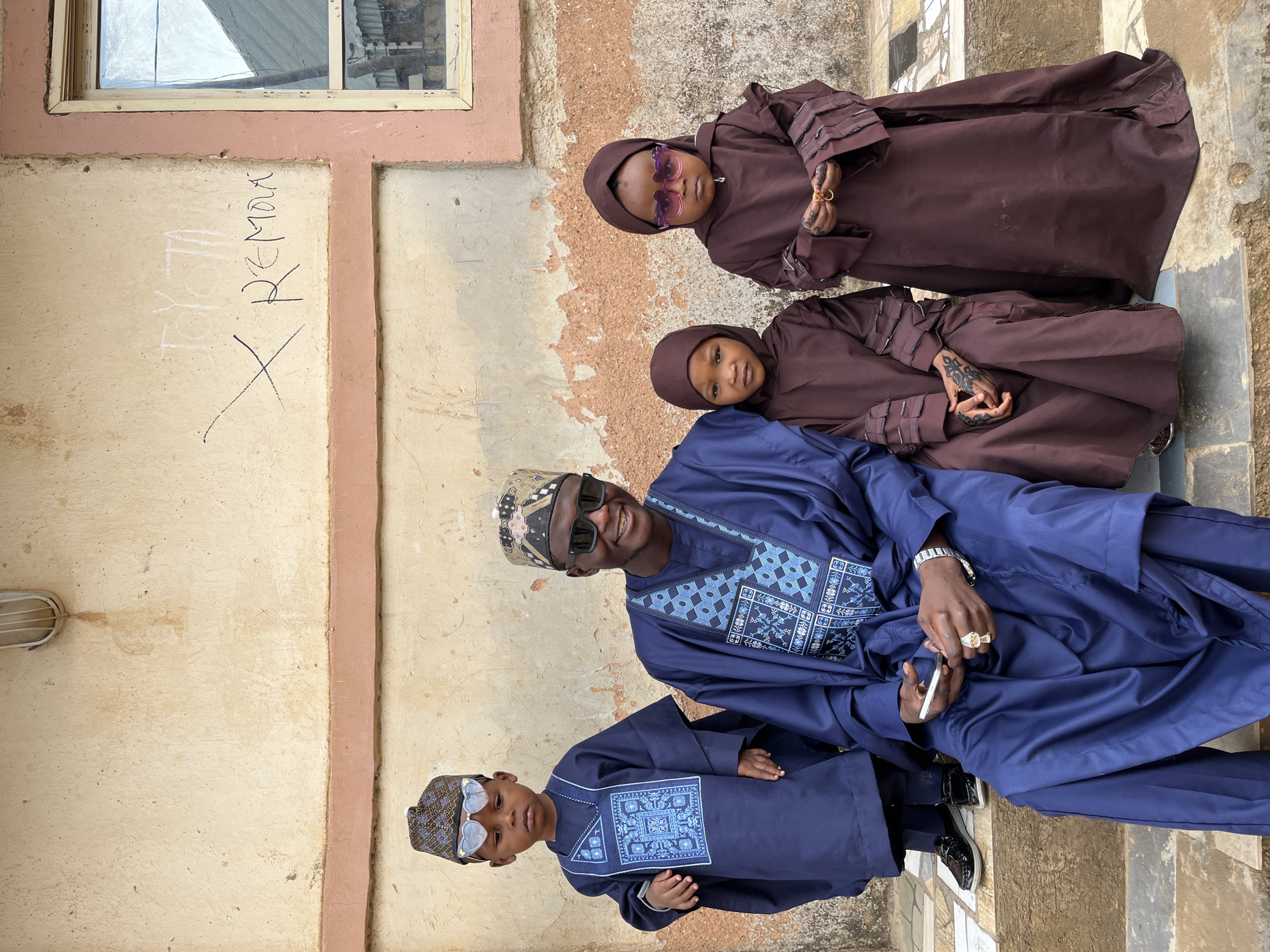 Four individuals stand or sit near a building with beige and reddish-brown concrete walls. The building has a window and some graffiti on the wall. The upper two individuals are children wearing brown hooded robes and sunglasses. They have traditional markings on their hands. Below them, an adult male in a blue patterned robe and hat sits and smiles, holding a white object. A young boy in a matching blue outfit stands in the foreground, also wearing sunglasses and a patterned hat. The scene appears to be outside on a tiled surface. The location is Jos, Nigeria.