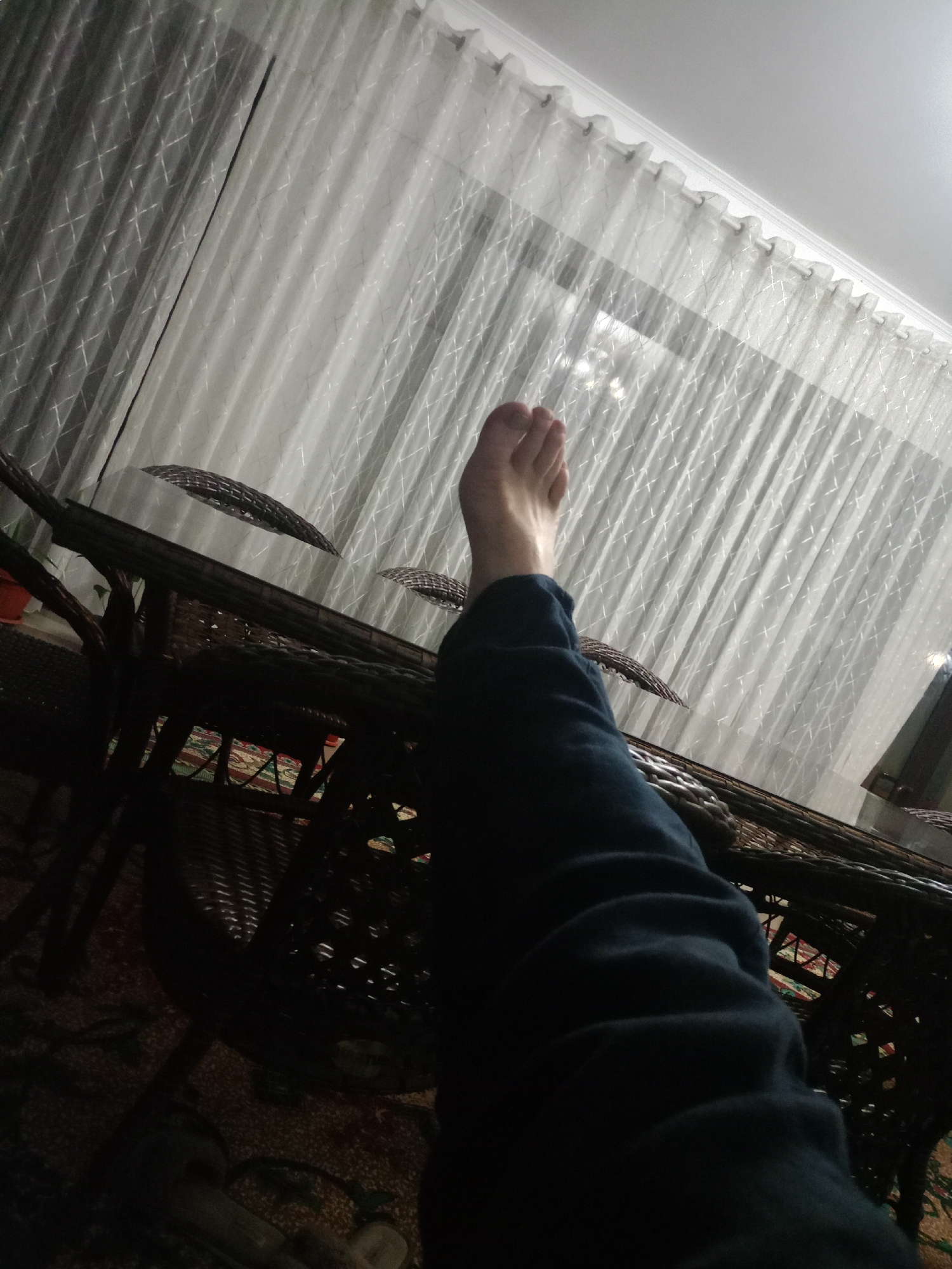 The image depicts a person's legs and feet extended towards the camera, resting on what appears to be a rattan table. The legs are covered by dark blue trousers. The background features sheer white curtains with a subtle geometric pattern, illuminated by natural light, suggesting a window. The setting is indoors, with the overall lighting dim except for the area near the curtains. The floor is covered by a patterned rug, partially visible beneath the table. The overall impression is one of relaxation or casual repose in a domestic environment. The image does not provide specific information to determine its location in Kandauraul, Russia.