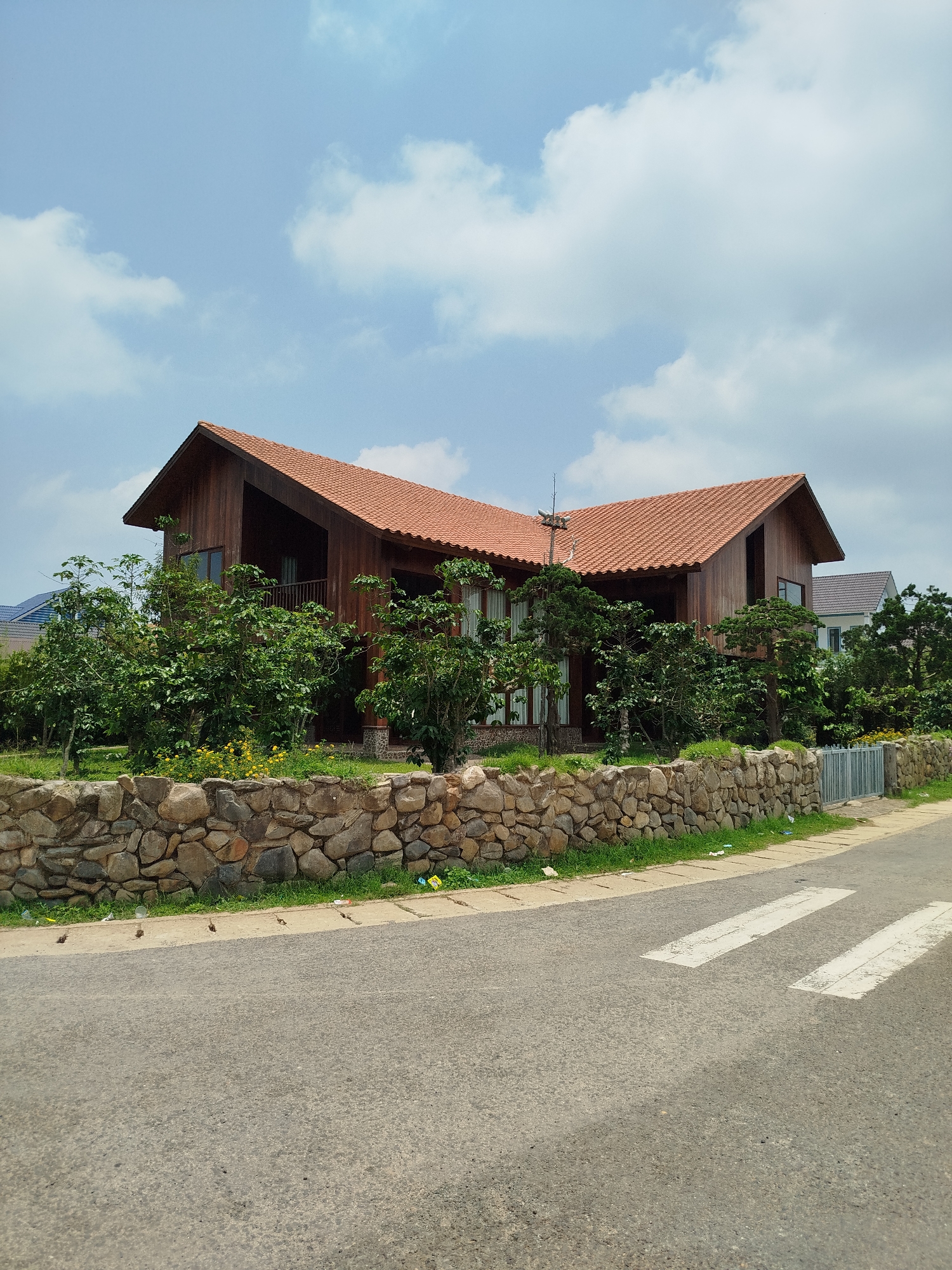 The image shows a wooden house with a terracotta tiled roof in Đa Nghịt, Vietnam. The house is set back from the road and fronted by a stone wall and greenery. The road has a pedestrian crossing marked on it. The sky is blue with scattered clouds. Another house is visible in the background.