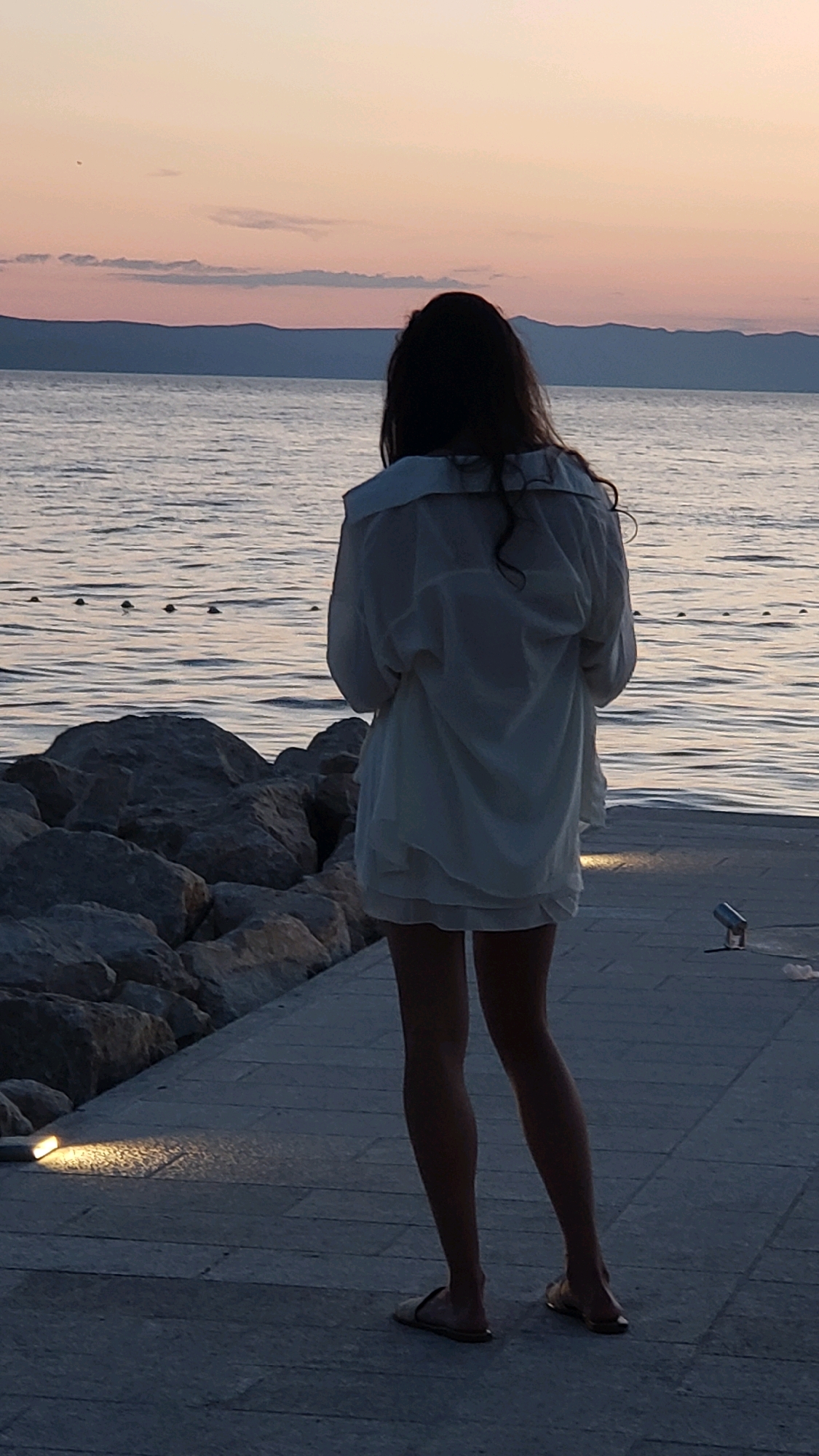 A person stands on a paved walkway next to a rocky shoreline, facing a body of water. The person is viewed from behind, wearing a white, loose-fitting garment and sandals. The sky is a dusky pink and orange, indicating either sunrise or sunset. The water is calm, with a few buoys visible on the surface. In the background, a hazy mountain range is visible across the water. The walkway is illuminated by discreet ground lights. The overall impression is one of a tranquil seaside scene. The location is Općina Tučepi, Croatia.
