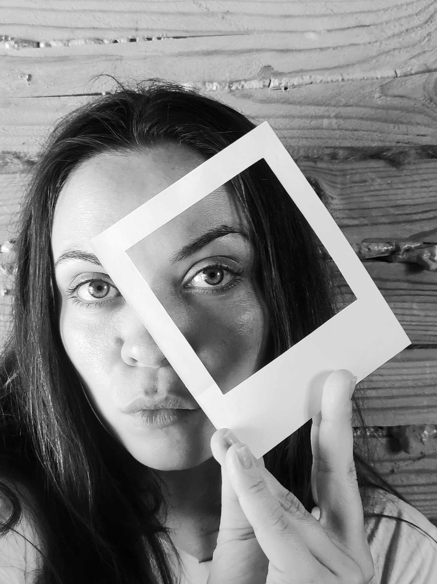 A grayscale, close-up, chest-up portrait captures an adult female against a horizontal wooden plank background, location in the city of Unknown, Unknown. The subject, with long, dark hair parted centrally, looks directly at the viewer with a slightly pouting expression. She holds a white instant photograph frame in her right hand, angled across her face. The frame partially obscures the right side of her face (viewer's left) and her nose, while clearly framing her left eye (viewer's right). Her right hand's fingers are visible, gripping the bottom right edge of the frame. The background consists of rough-hewn, horizontally oriented wooden planks with visible grain and texture.