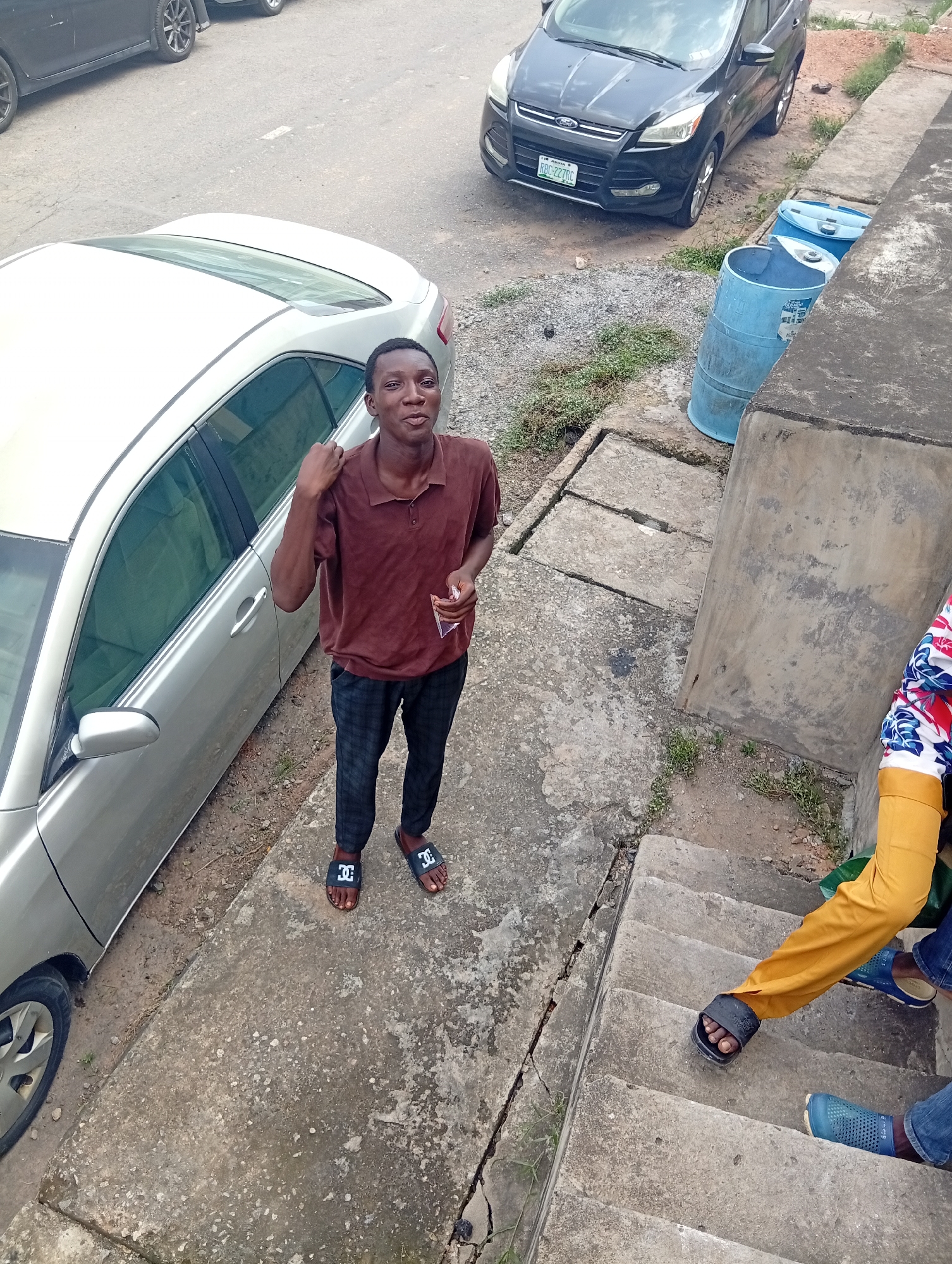 The image captures a man in Shangisha, Nigeria, standing outdoors next to a silver car. He wears a maroon polo shirt, checkered pants, and black sandals. In the background, a black SUV is parked along the street, next to two blue barrels near a concrete wall. Steps lead down from the wall, where another person wearing a patterned shirt and yellow pants stands. The environment appears to be a street-side or parking area with concrete surfaces and some vegetation.