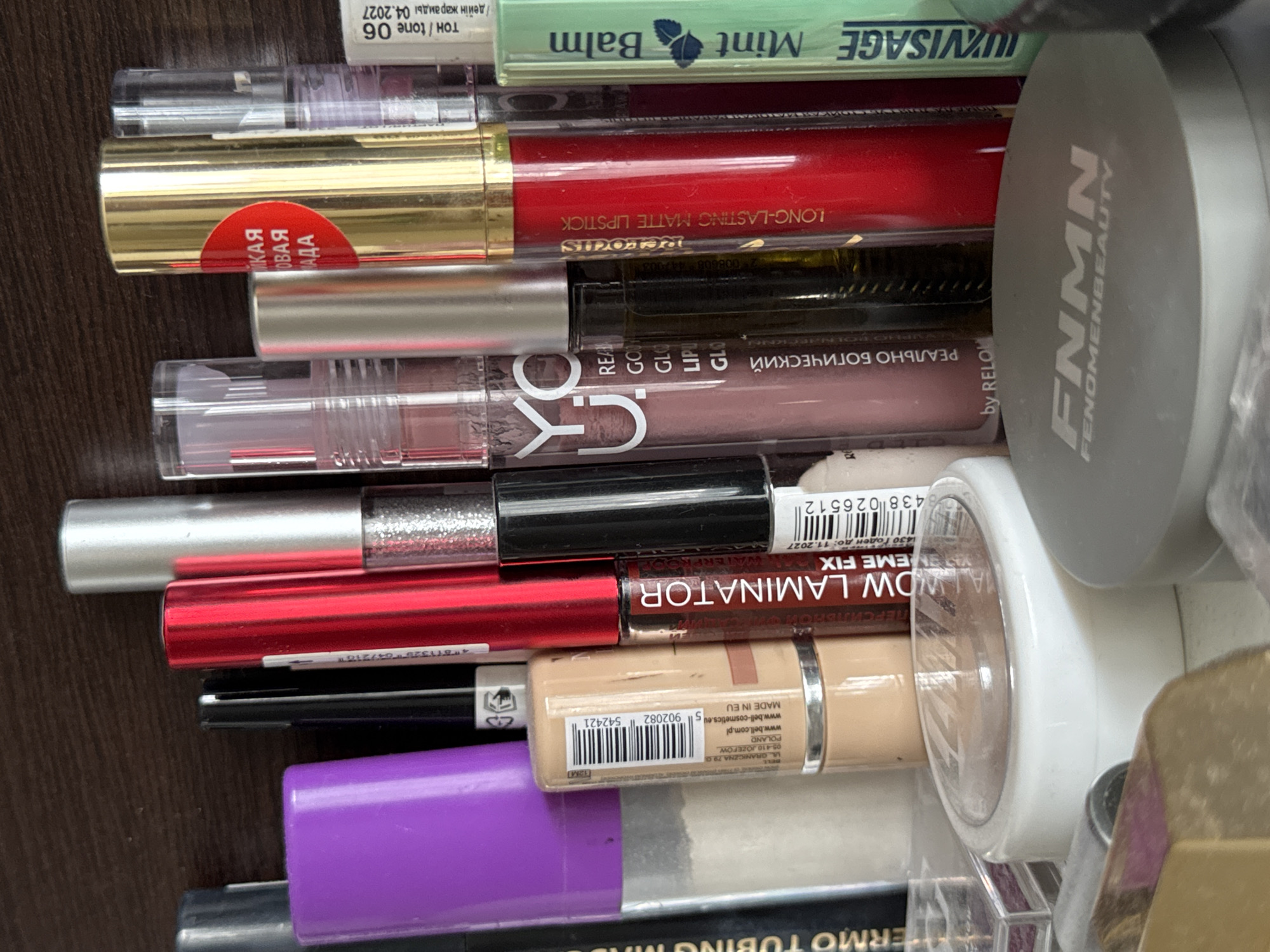 The image shows a close-up, eye-level view of a collection of makeup products, primarily lipsticks and liquid lipsticks, arranged vertically. The products are on a dark, wood-grain surface. From left to right, the visible items include a purple-capped tube, a beige foundation stick with a barcode and "MADE IN EU" label, a red-capped lipstick tube, a black tube with a silver cap, a silver-capped lipstick, a clear tube with a pinkish-brown liquid and white text "C:YO", a clear tube with a dark liquid and silver cap labeled "REVOUS", a bright red-capped lipstick labeled "LONG-LASTING MATTE LIPSTICK", a metallic gold tube, a clear tube with a pale liquid, and a green tube labeled "LVISAGE Mint Balm". In the foreground, a round, white makeup compact with a clear lid is visible, and behind it, a larger, grey makeup compact with the logo "FNMN FENOMENBEAUTY". The overall impression is of a makeup collection organized on a shelf or vanity. The context of Яромінскі сельскі Савет, Belarus is not visually evident from the image itself.