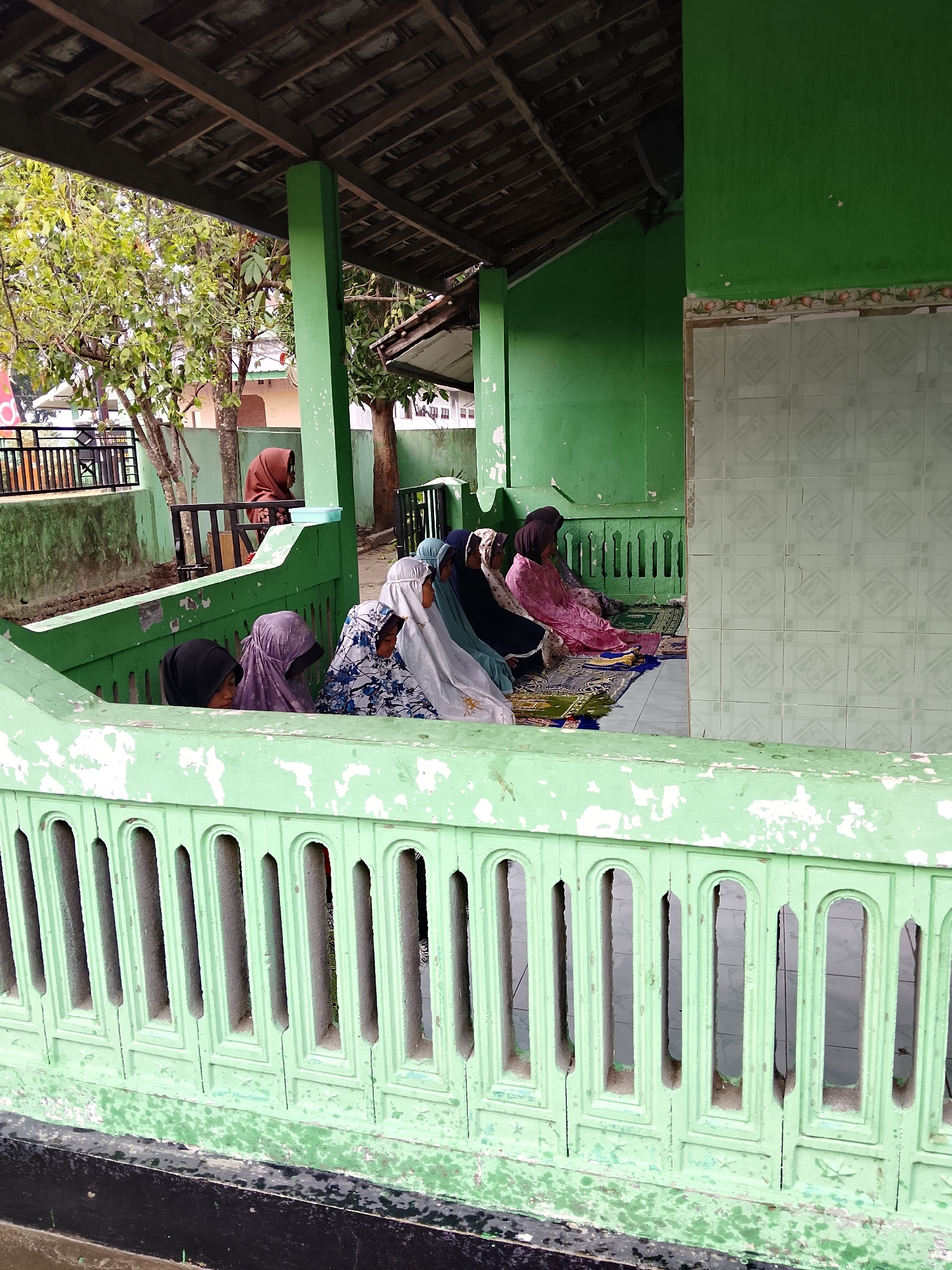 In Kepuh, Indonesia, a group of Muslim women are gathered, seated on prayer mats in what appears to be an outdoor shaded structure with a tiled back wall. The structure is painted green, and a green decorative railing is in the foreground. The women are wearing hijabs of various colors, including black, purple, white with a floral pattern, and pink. Some are looking down, seemingly in prayer or contemplation. The environment suggests a religious or communal gathering space.